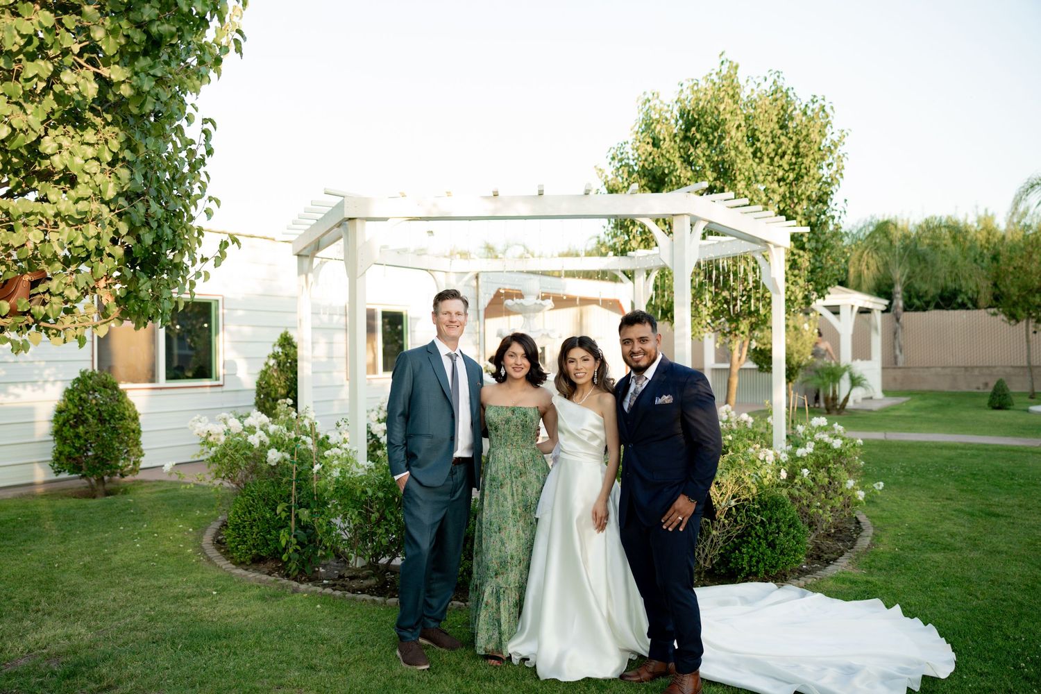 Wedding party of four posing in front of a white pergola on a lush green lawn.
