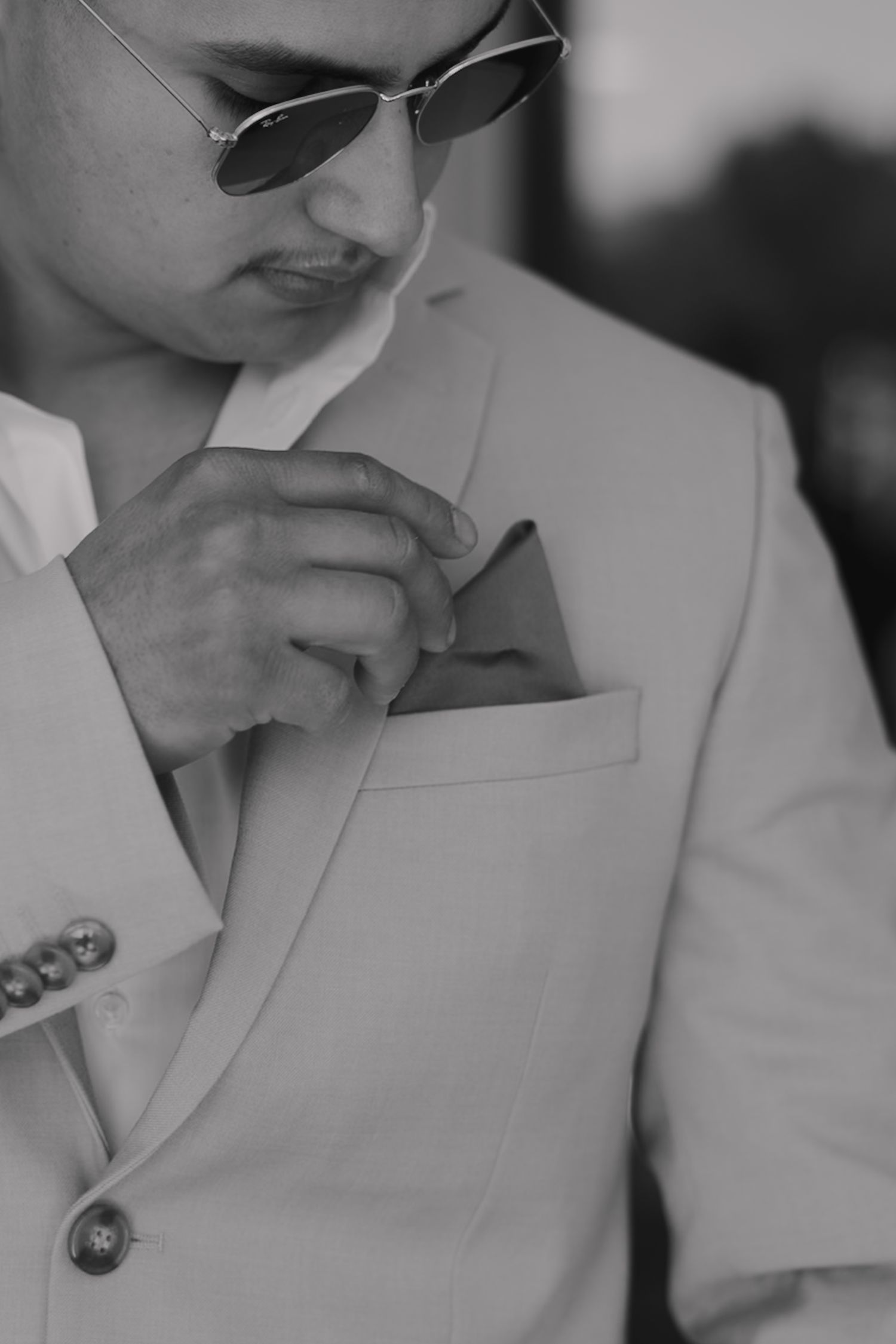 Stylish groom in light suit adjusting pocket square wearing aviator sunglasses, black and white photo.