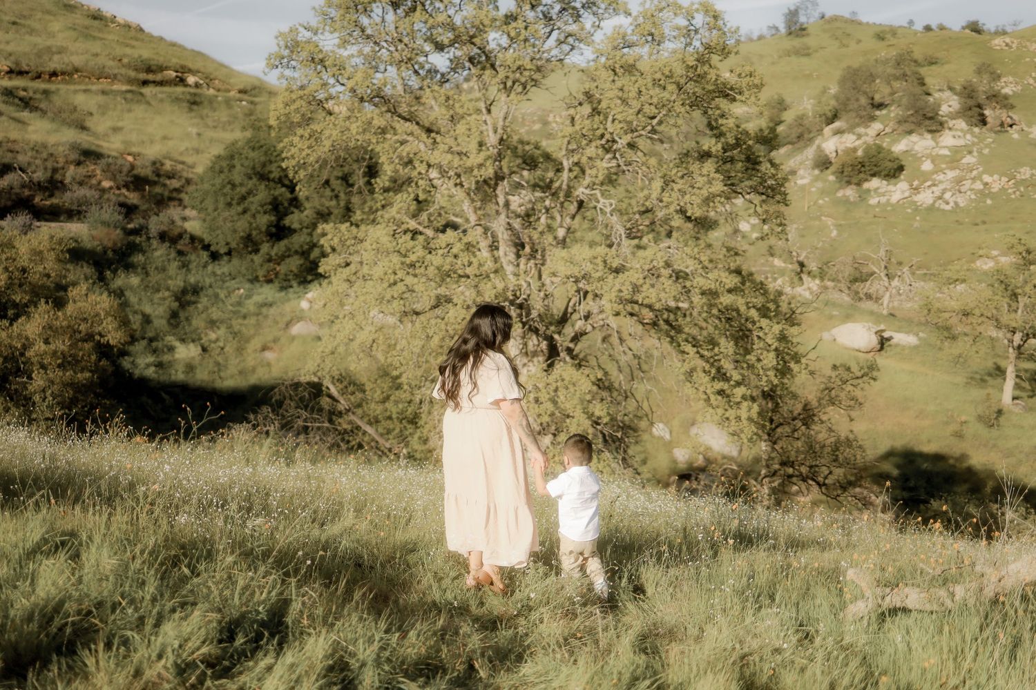 A couple in light colored clothing walks through a scenic hillside meadow during golden hour.