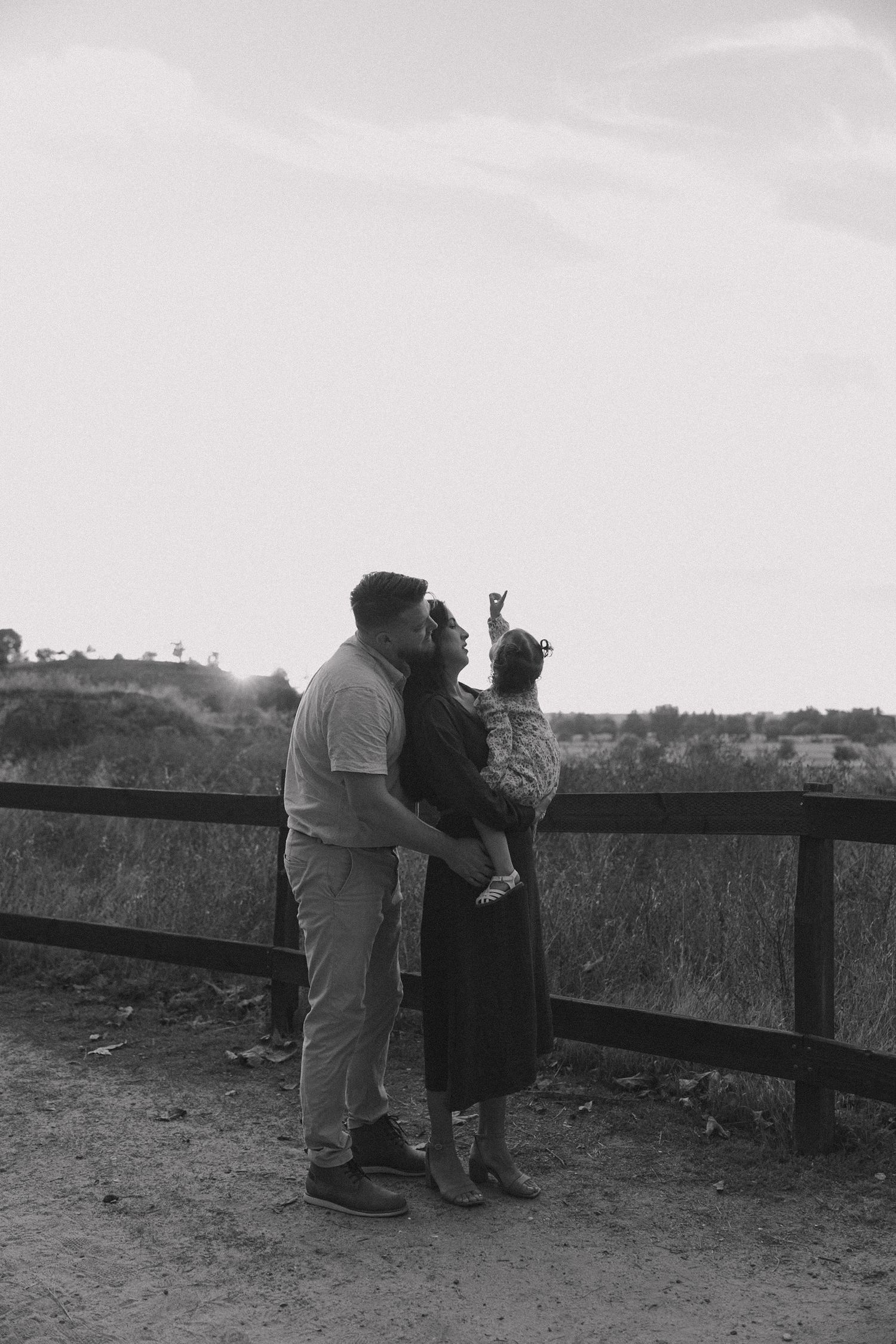 Two people lean over a wooden railing to look at something in the distance in this black and white photograph.