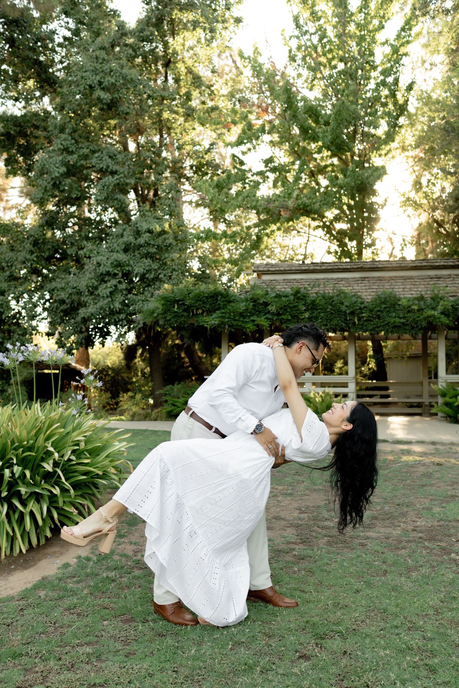 A romantic dip pose between a couple in white attire surrounded by lush garden scenery.