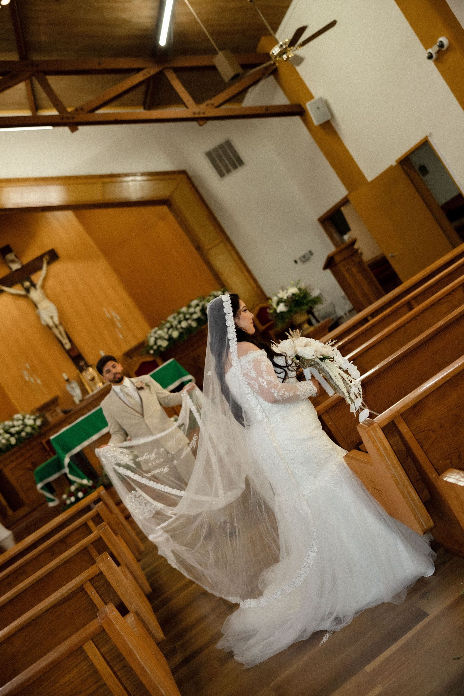 Bride in white gown and veil walks church aisle toward groom during wedding ceremony with wooden pews and cross.