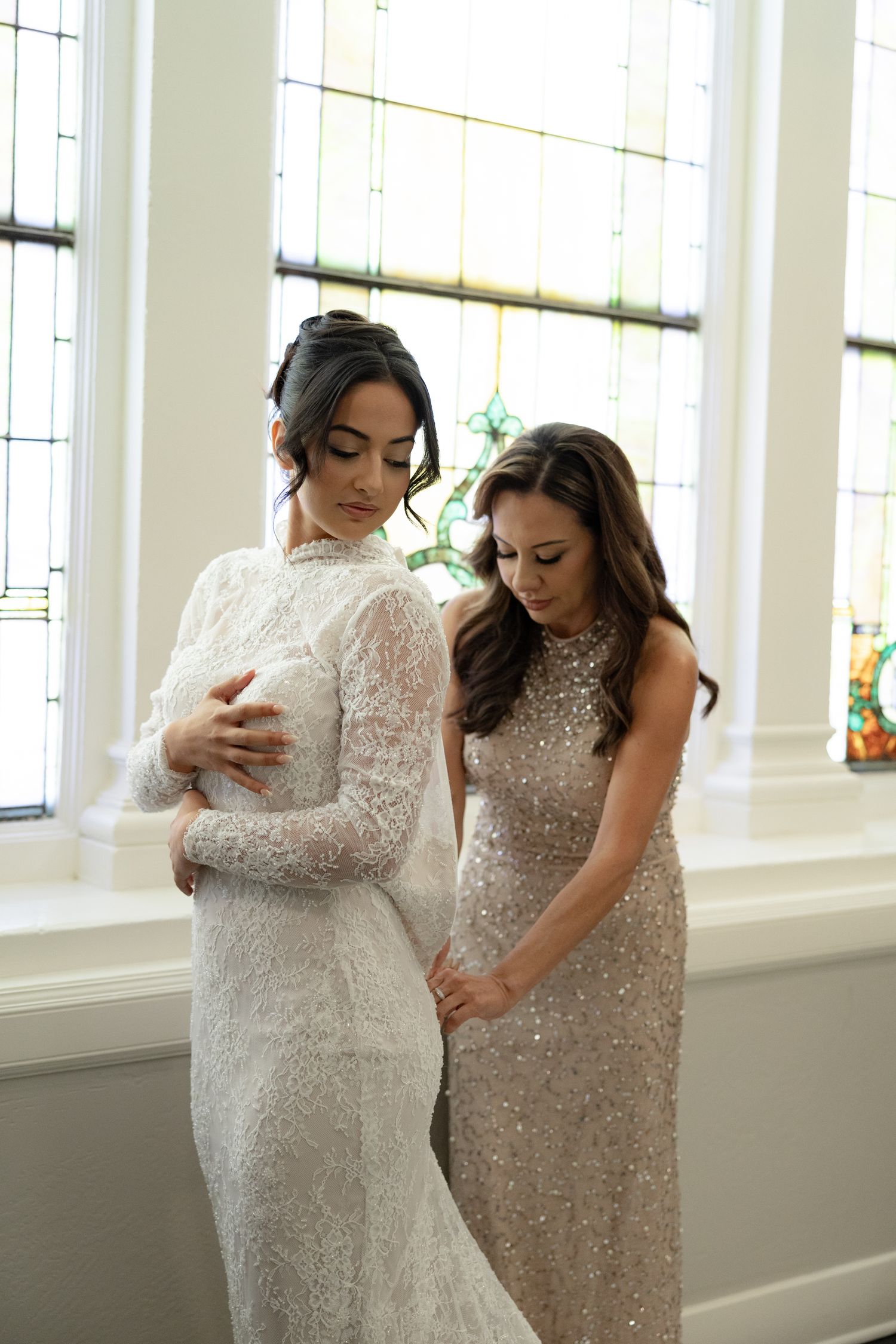 Bridesmaid in sequin gown helps bride in lace wedding dress near stained glass windows.