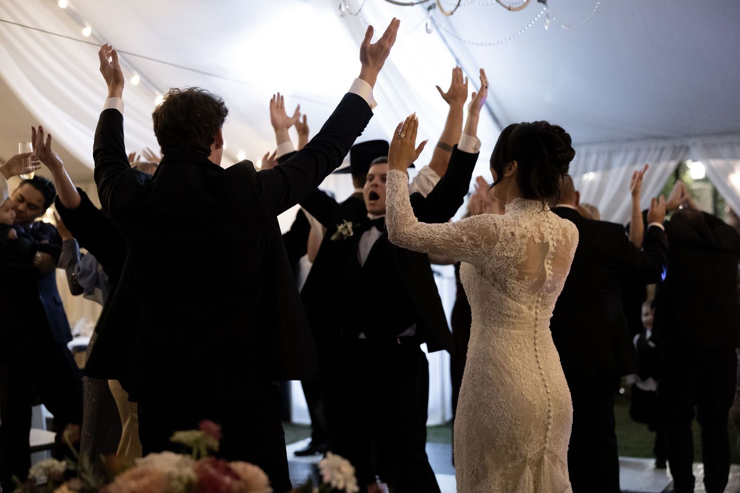 Bride and groom celebrate with hands raised surrounded by cheering guests at wedding reception.