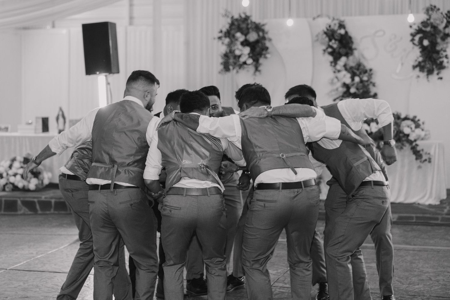 Groomsmen in gray suits huddle together in a group embrace at a wedding reception venue in black and white photo.