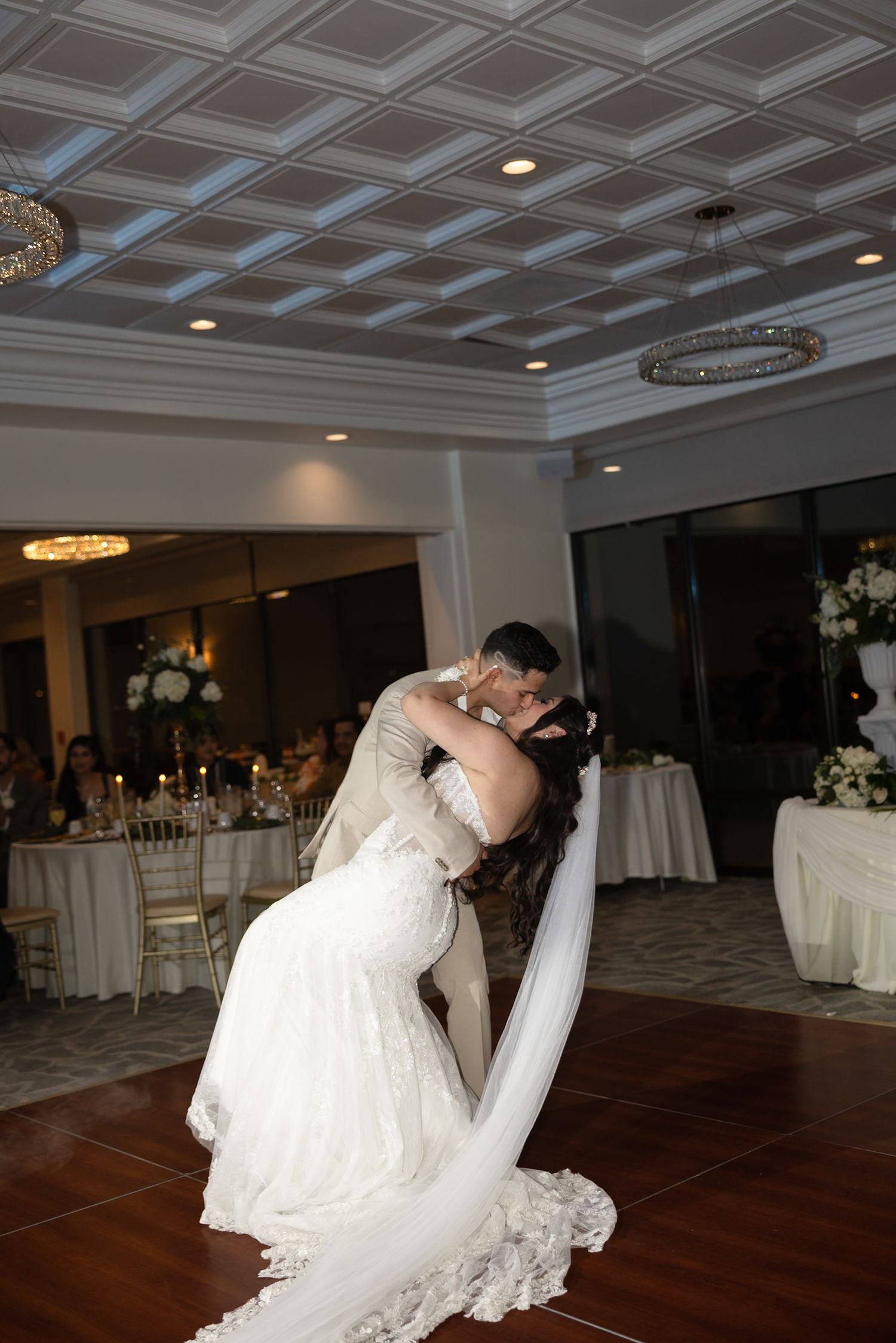 Groom dips bride for romantic first dance kiss in elegant ballroom with guests and candlelit tables behind.