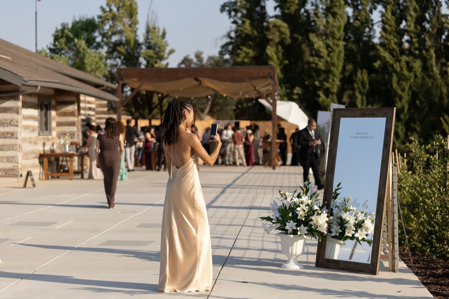 A woman in a champagne gown stands near a floral arrangement and mirror at an outdoor wedding venue.