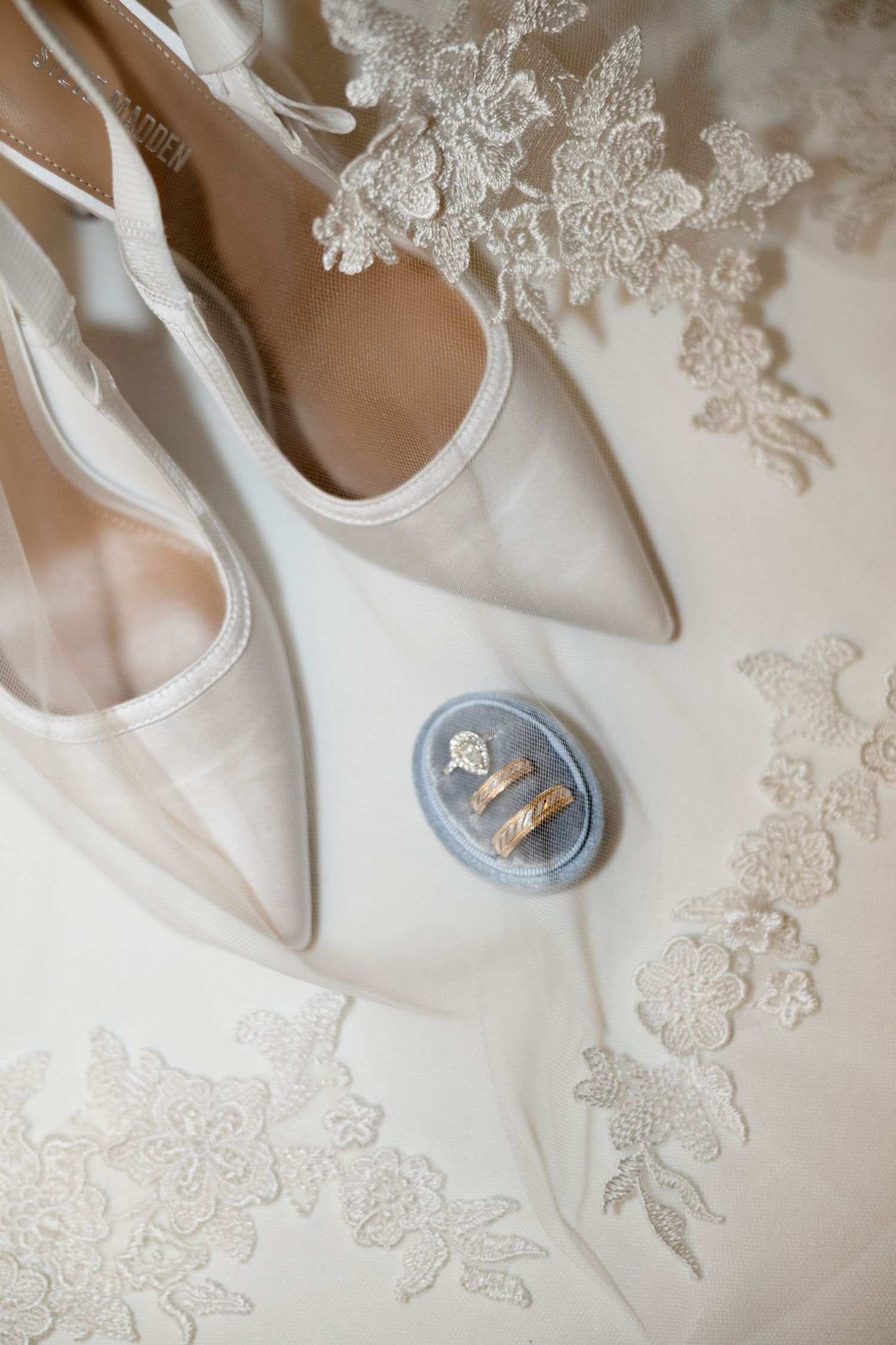 Elegant bridal flat lay featuring white satin heels, gold wedding rings on blue velvet, and delicate lace veil details.