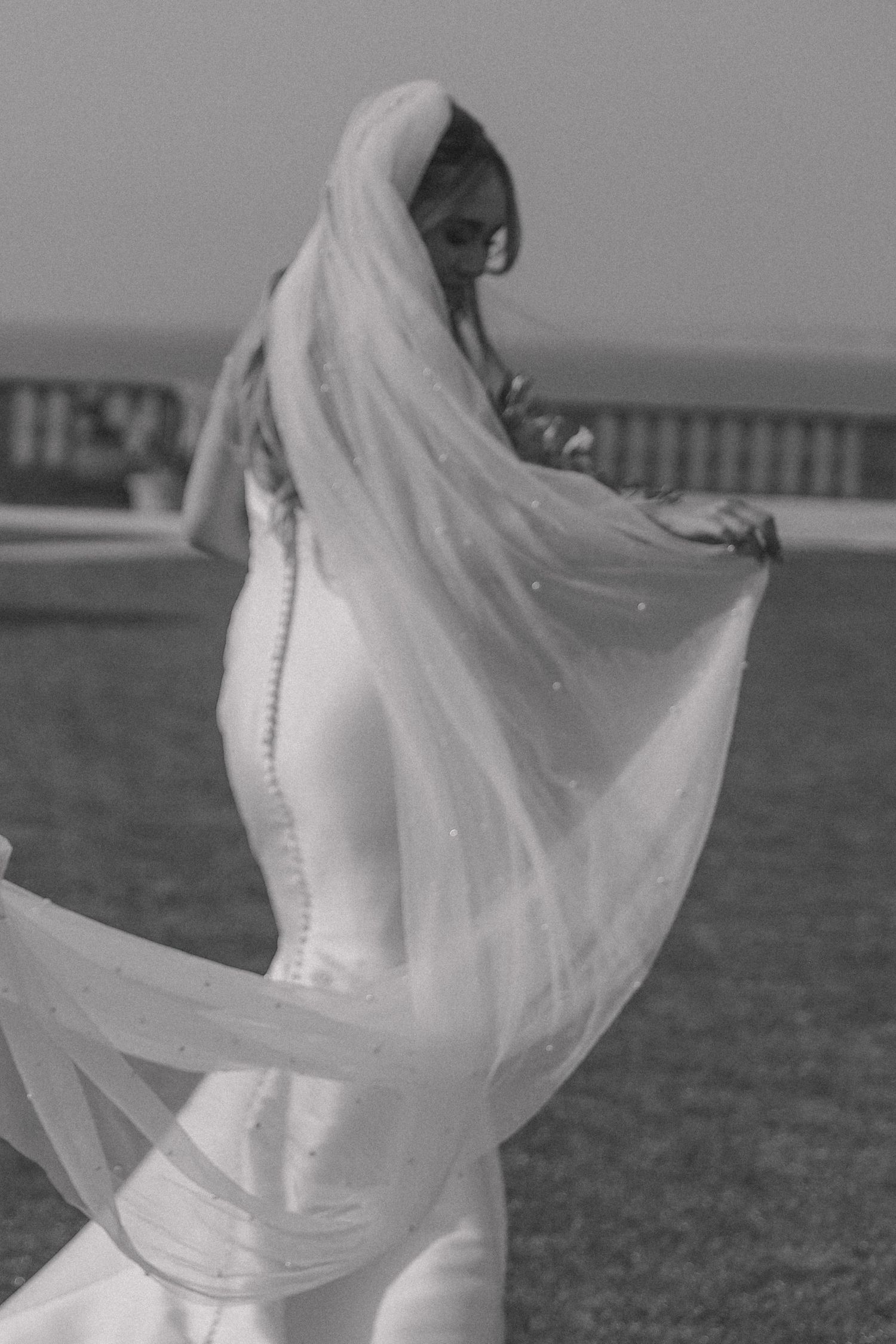 A flowing white wedding dress veil billows in the wind against a scenic outdoor backdrop.