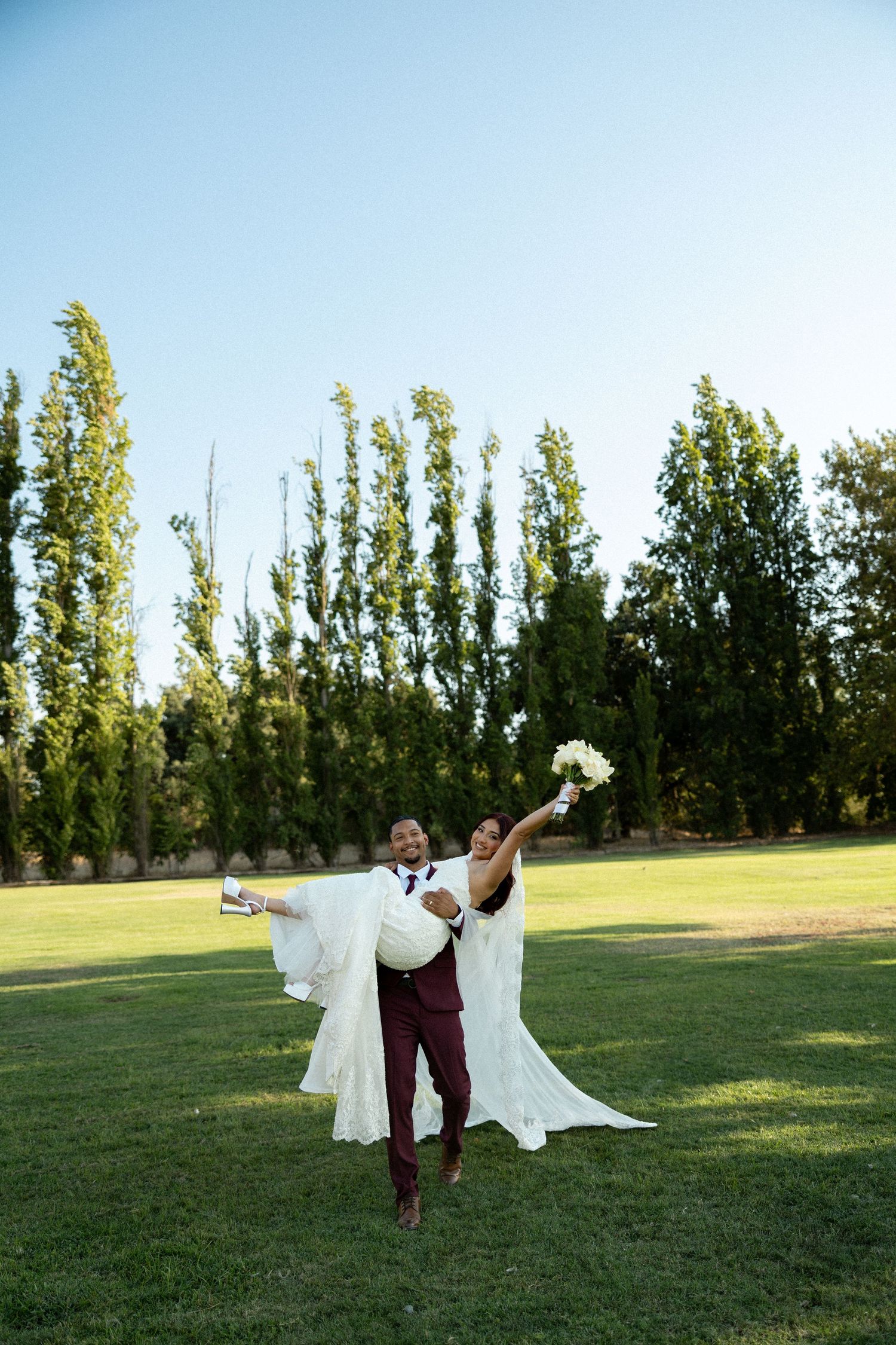 Groom in burgundy suit joyfully lifts bride in white wedding gown on sunny green lawn.