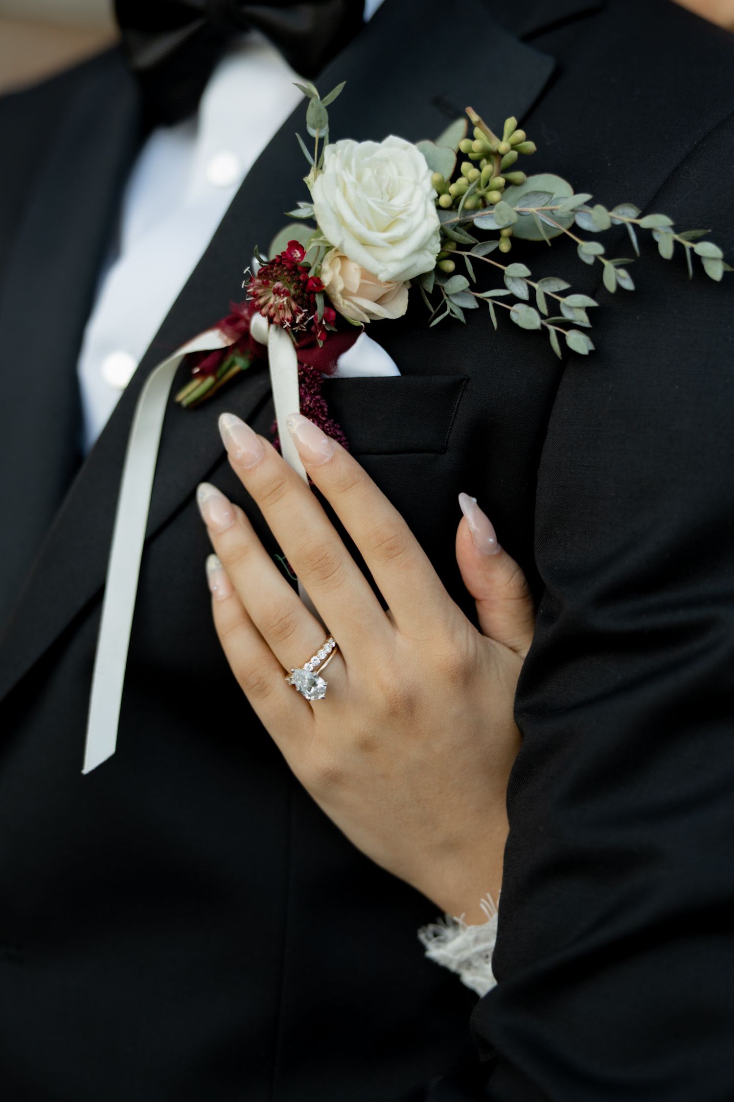 Close-up of bride's diamond ring and groom's white rose boutonniere on black tuxedo lapel.
