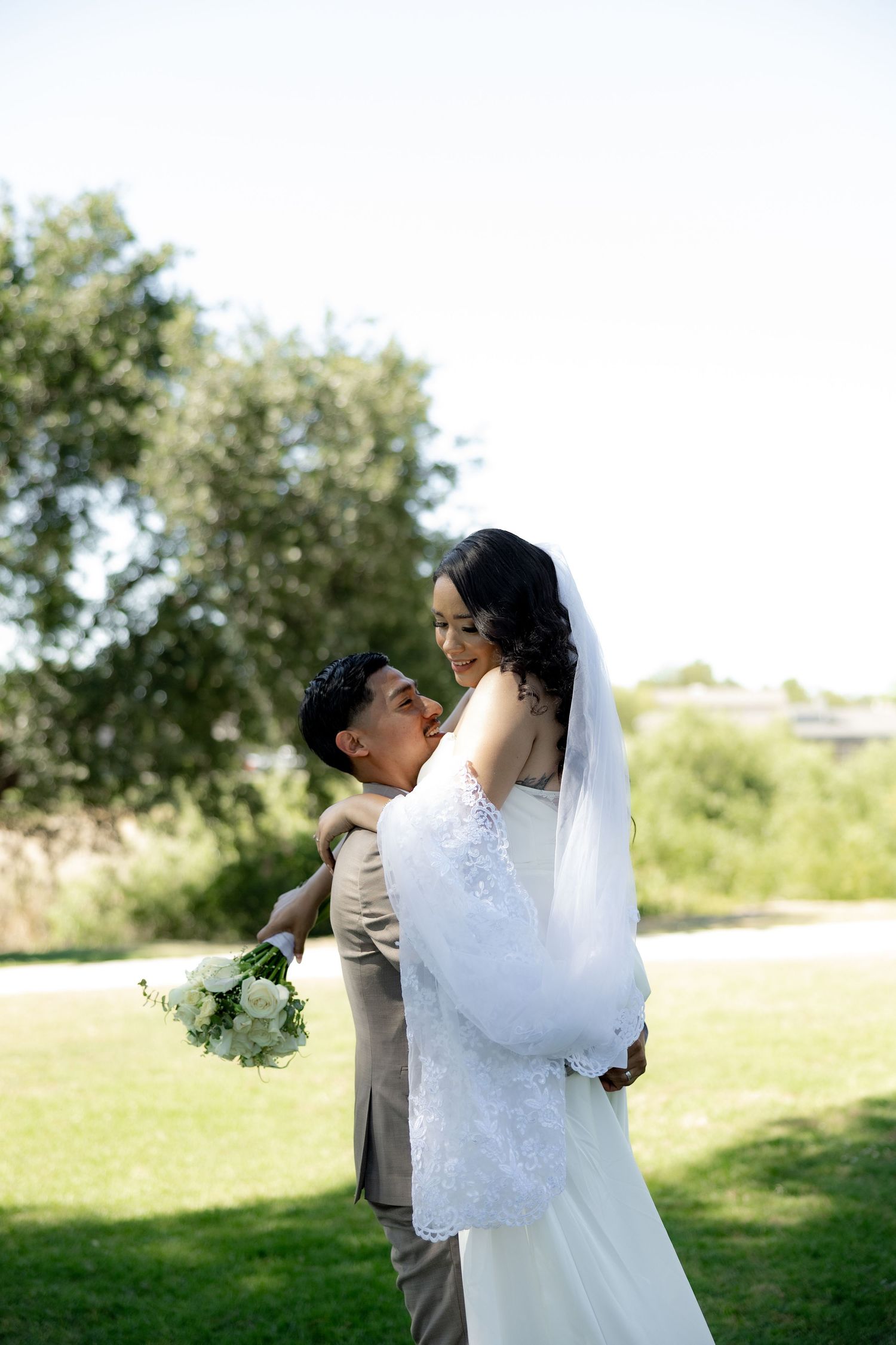 A groom in a tan suit lifts his bride in a white lace gown and veil outdoors on a sunny day.