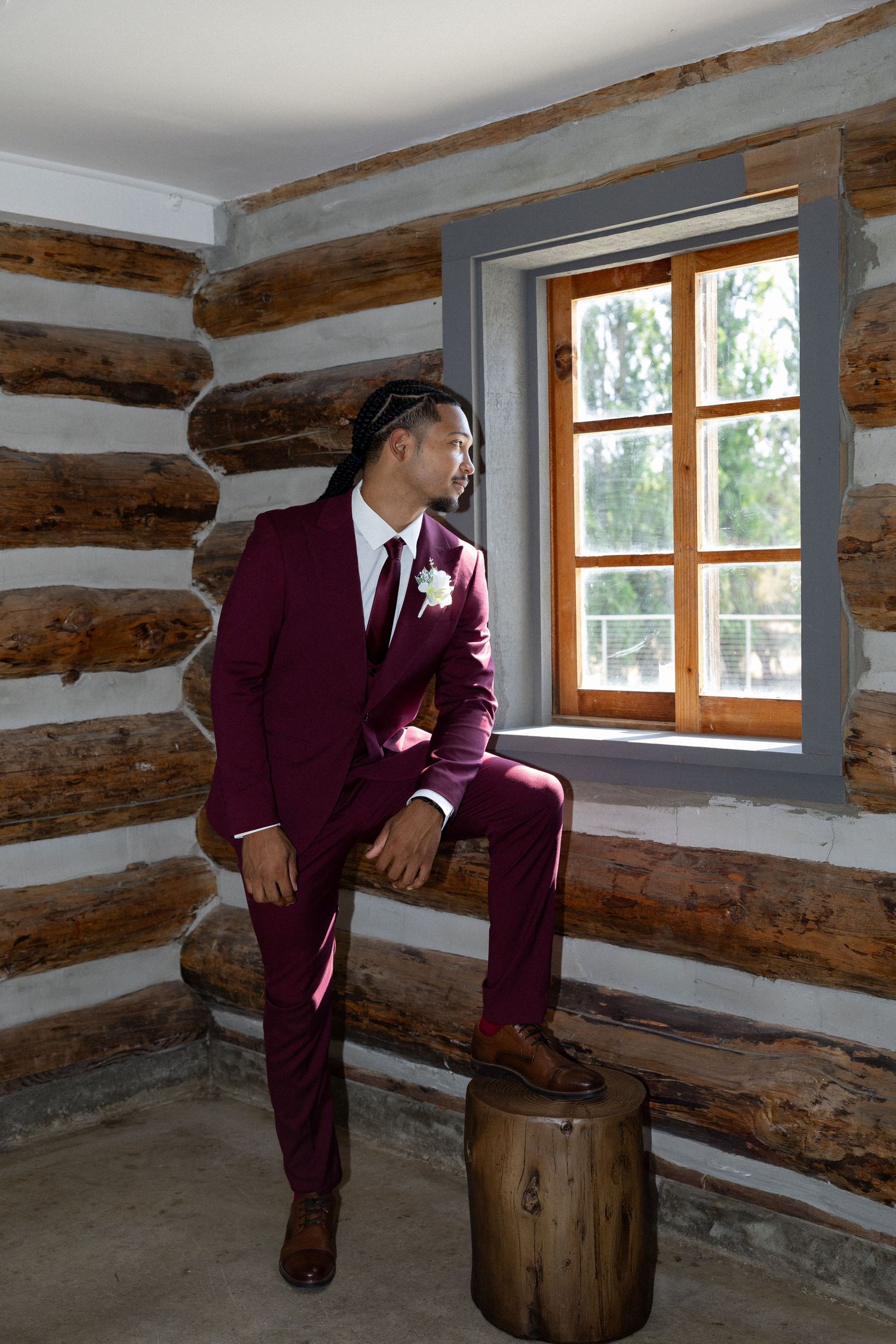 Groom in burgundy suit with boutonniere sits on wooden bench by window in rustic log cabin wedding venue.