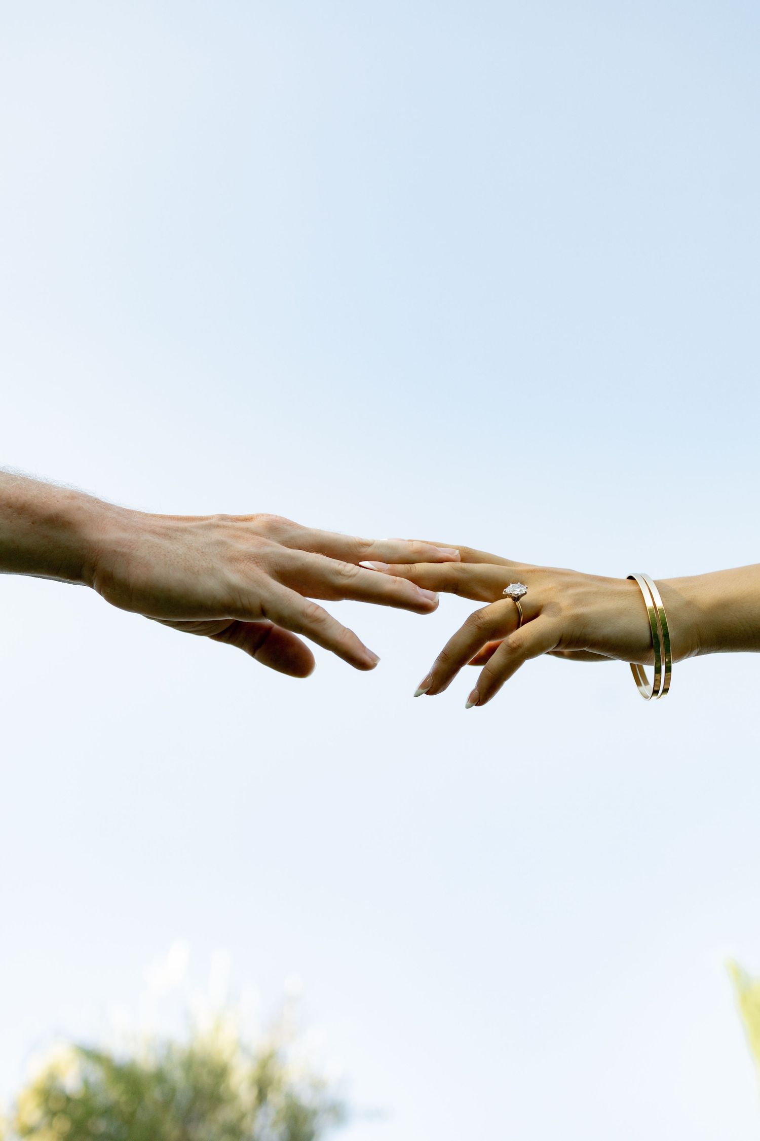 Two hands with wedding rings reaching towards each other delicately against a bright sky.