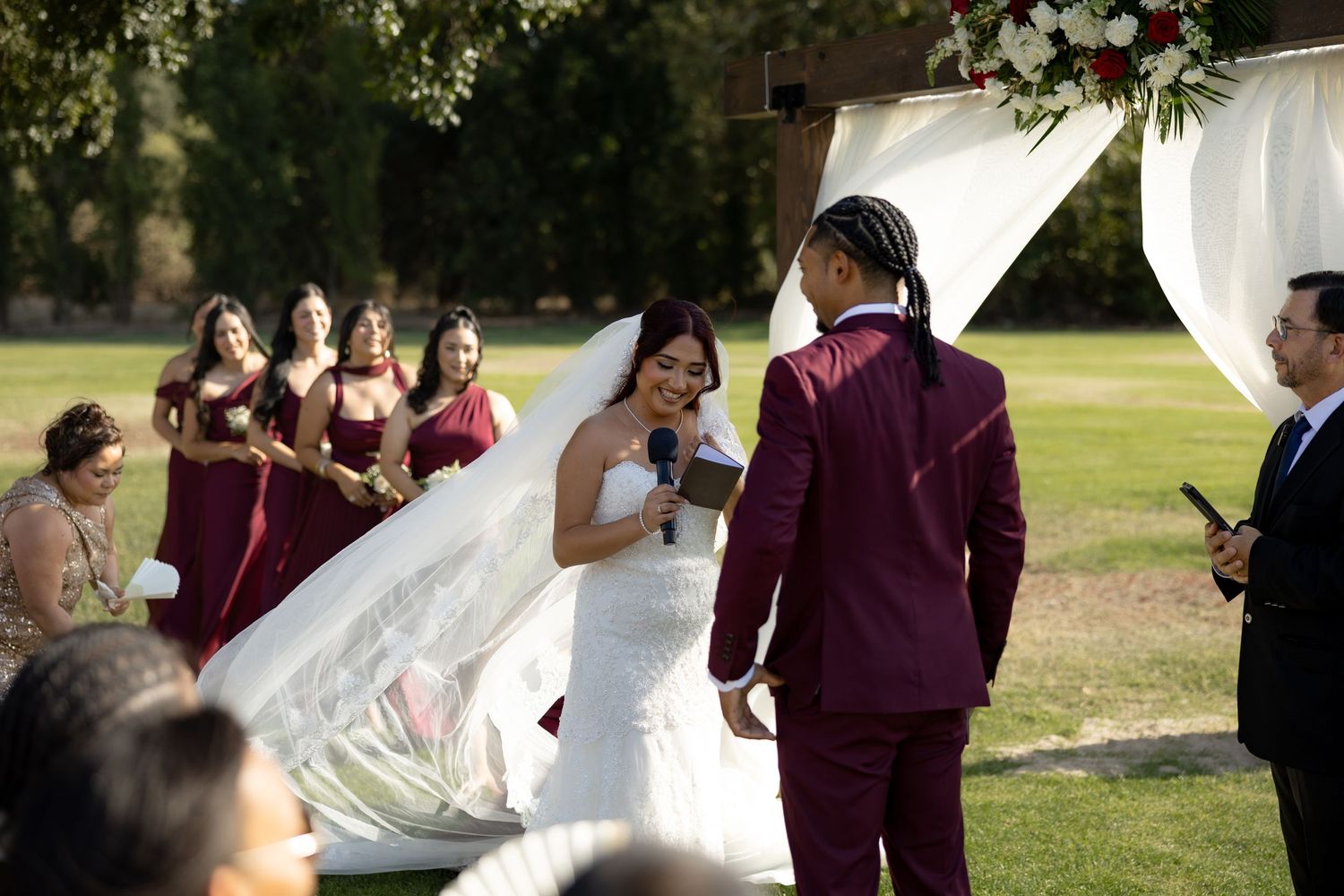 Bride in white gown reads vows to groom in burgundy suit at outdoor wedding ceremony with bridesmaids behind.