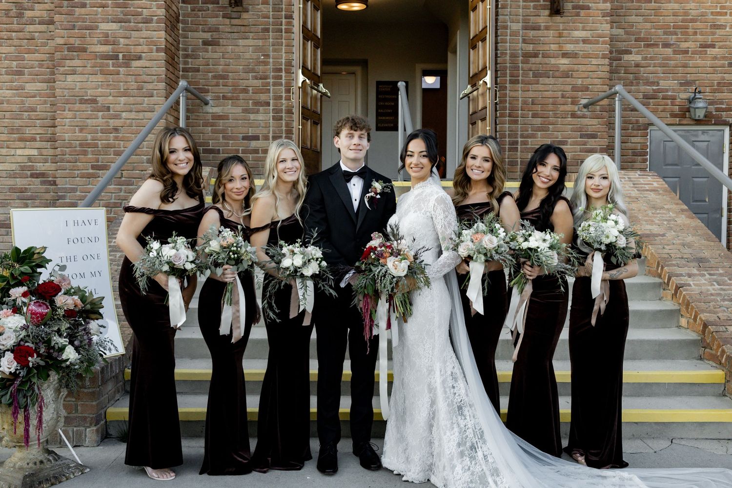 Wedding party in black velvet gowns and tuxedo poses with bride in white lace gown on church steps.