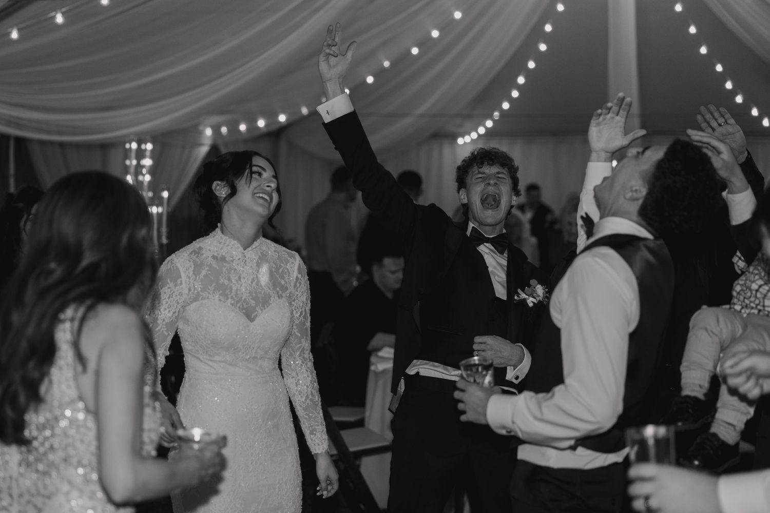 Black and white photo of a bride and groom dancing joyfully with guests at their wedding reception.