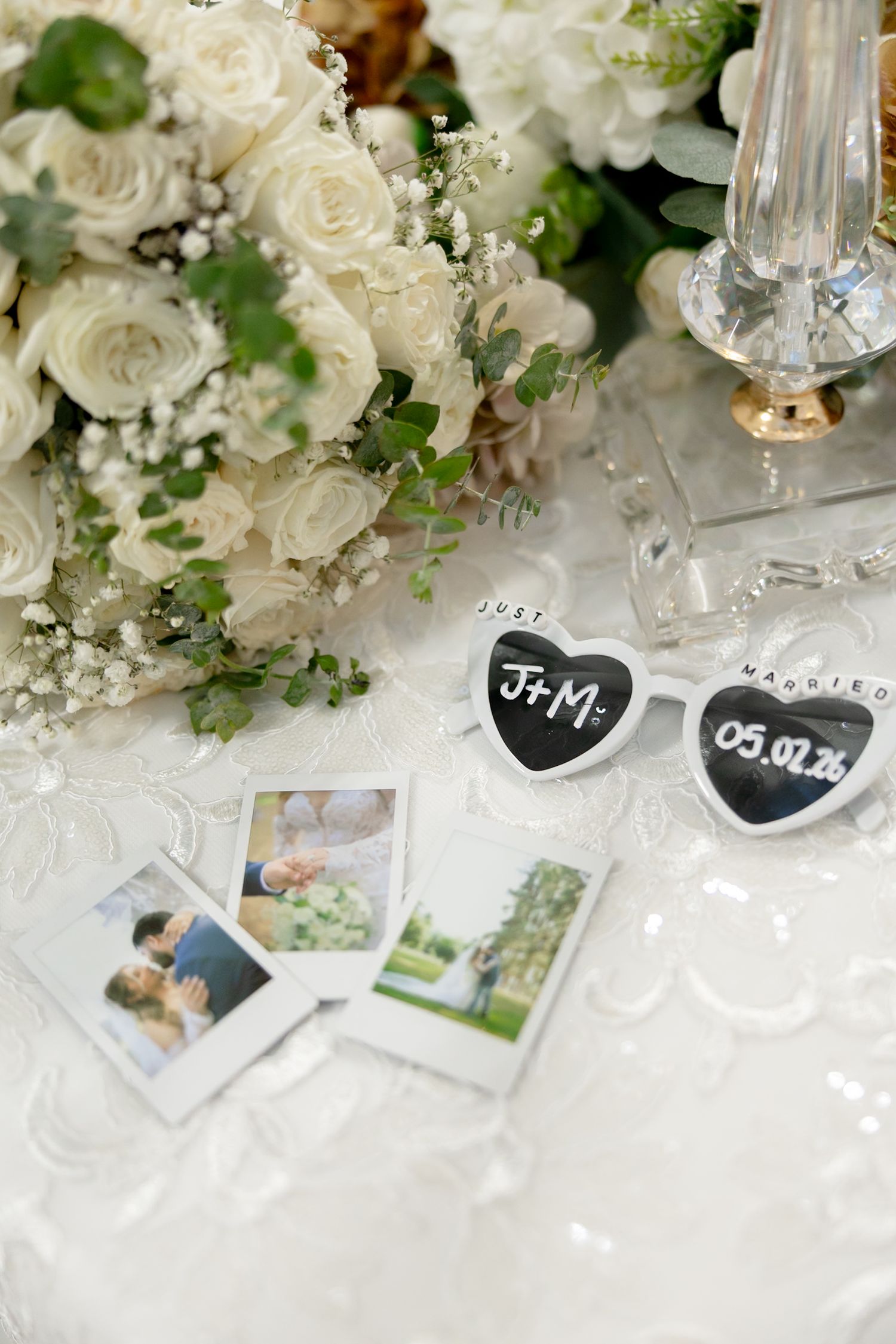 Wedding table with white rose bouquet, heart-shaped sunglasses reading J+M, Polaroid photos, and crystal decor on lace cloth.