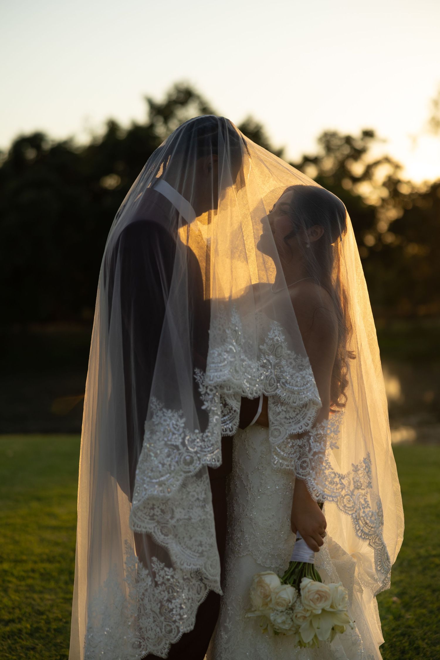 Bride and groom sharing a romantic kiss under a flowing lace wedding veil at golden hour outdoors.