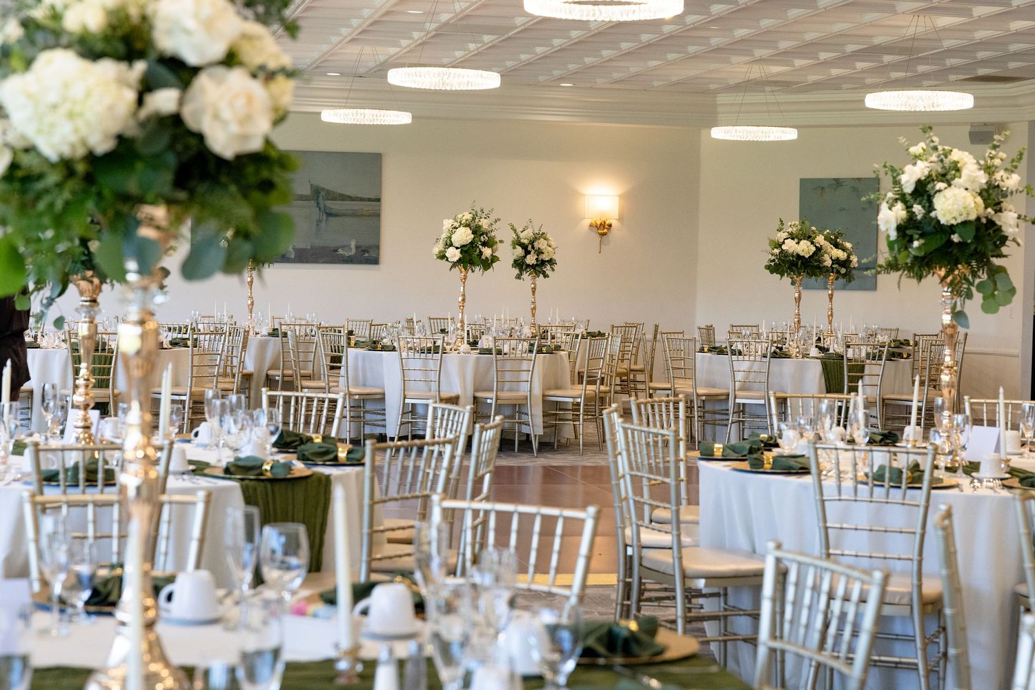 Elegant wedding reception hall with gold chiavari chairs, white floral centerpieces, and draped tables in a bright venue.