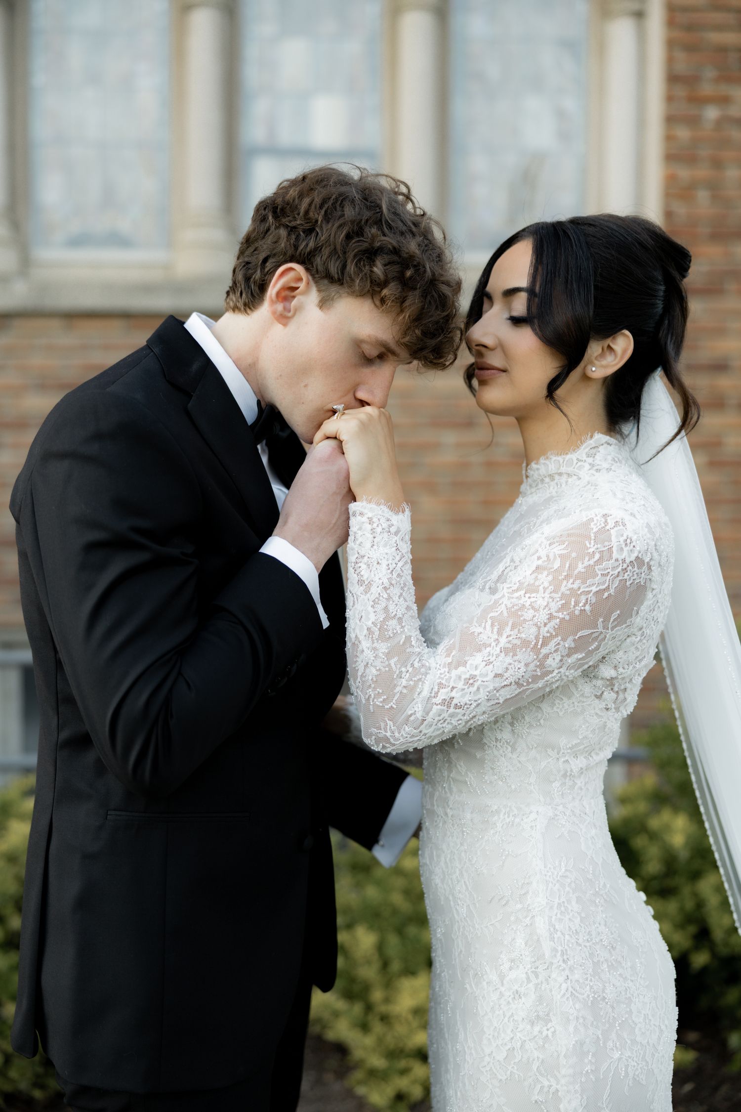 Groom in black tuxedo kissing brides hand as she smiles in lace wedding gown.