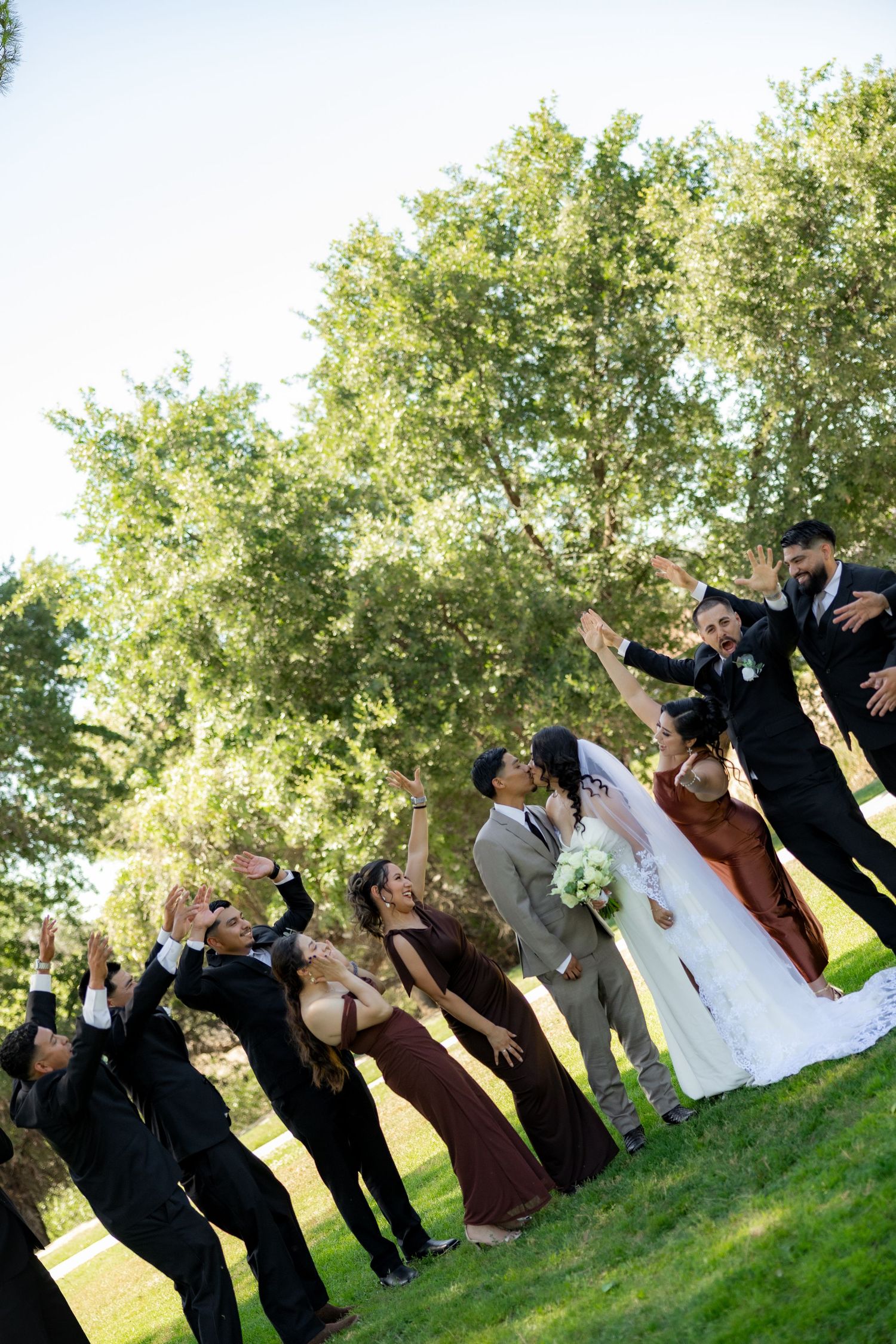 Wedding party celebrates outdoors as bride and groom kiss, surrounded by bridesmaids in burgundy and groomsmen in black suits.
