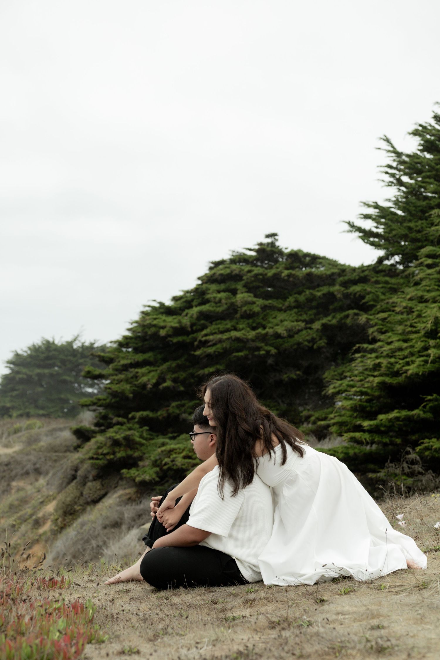 A person in a flowing white dress sits on a coastal cliff overlooking evergreen trees on a misty day.