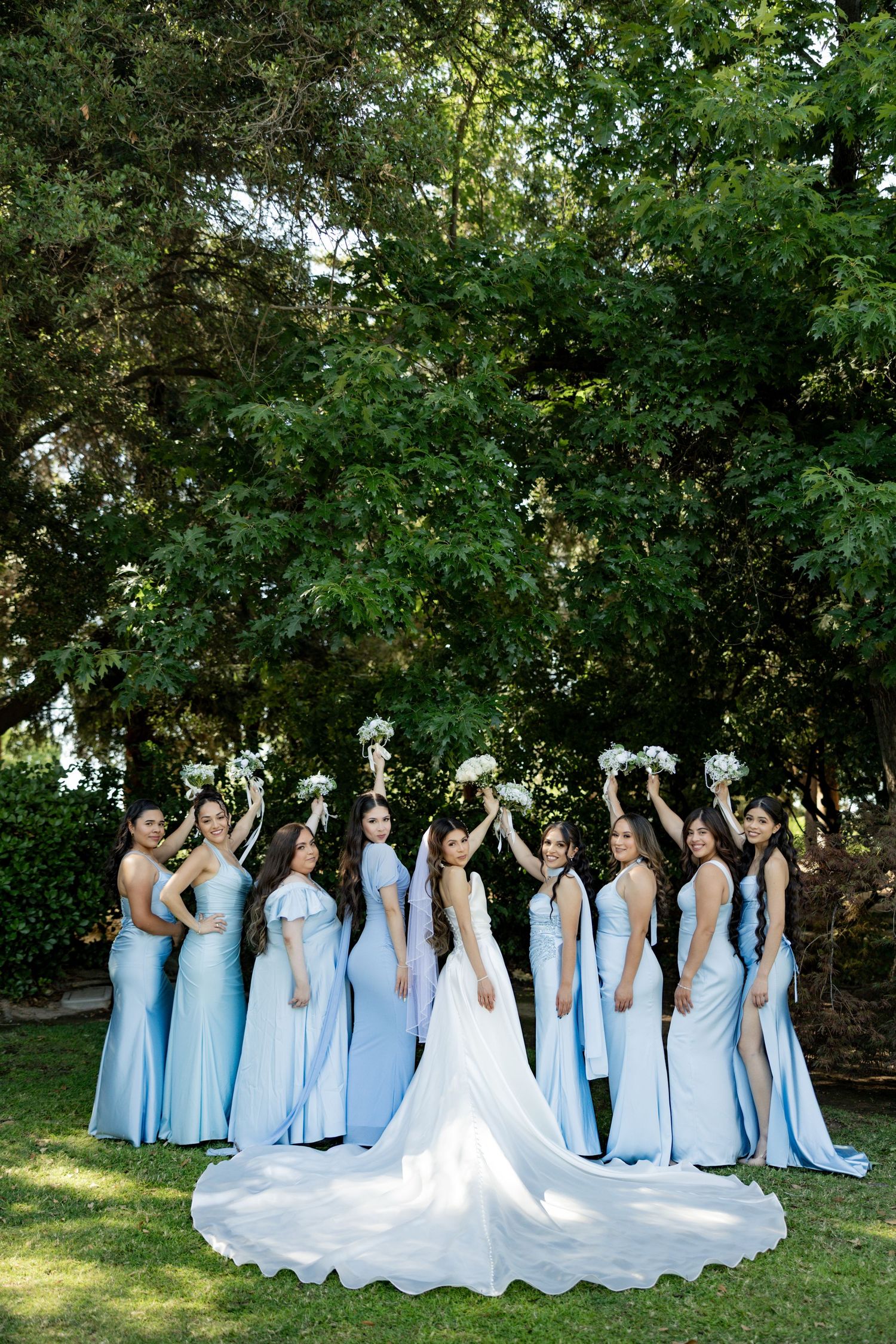 Bride in white gown with bridesmaids in light blue dresses raising bouquets outdoors among lush green trees.