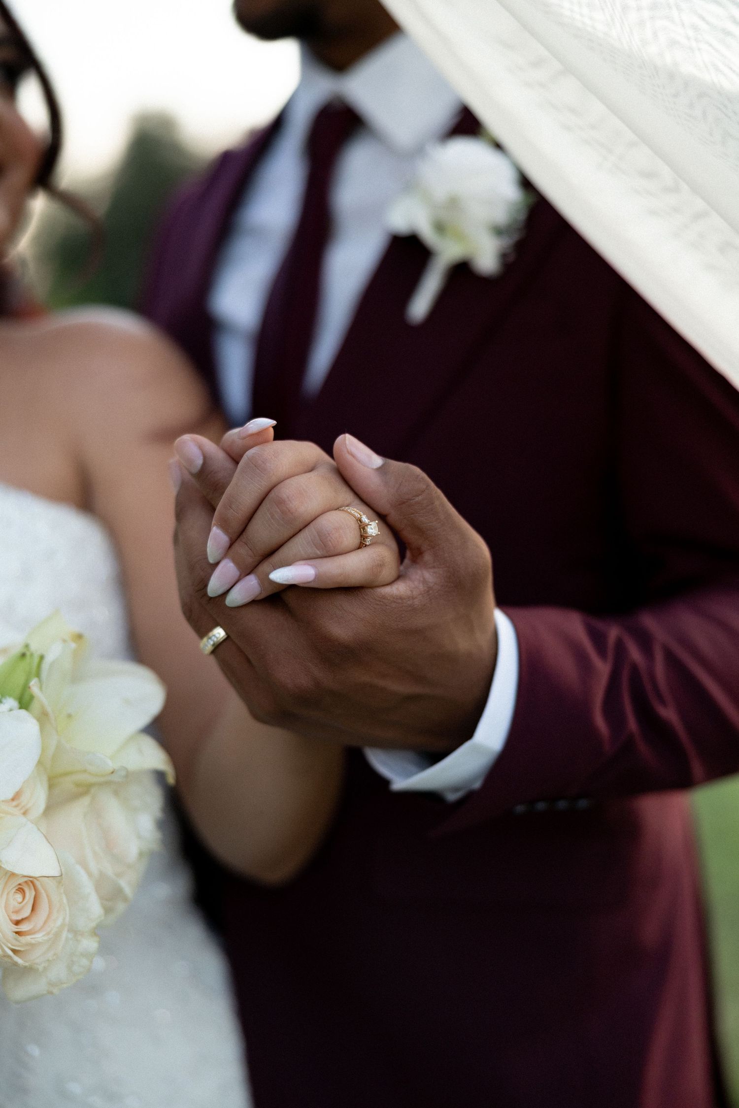 Bride and groom holding hands at wedding, bride wearing gold ring with white manicure, groom in burgundy suit holding bridal bouquet.