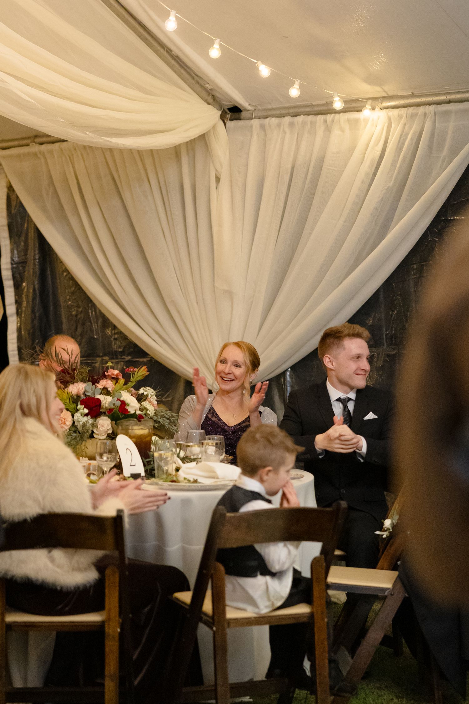 Wedding guests laughing and clapping at a reception table under a draped tent with string lights.