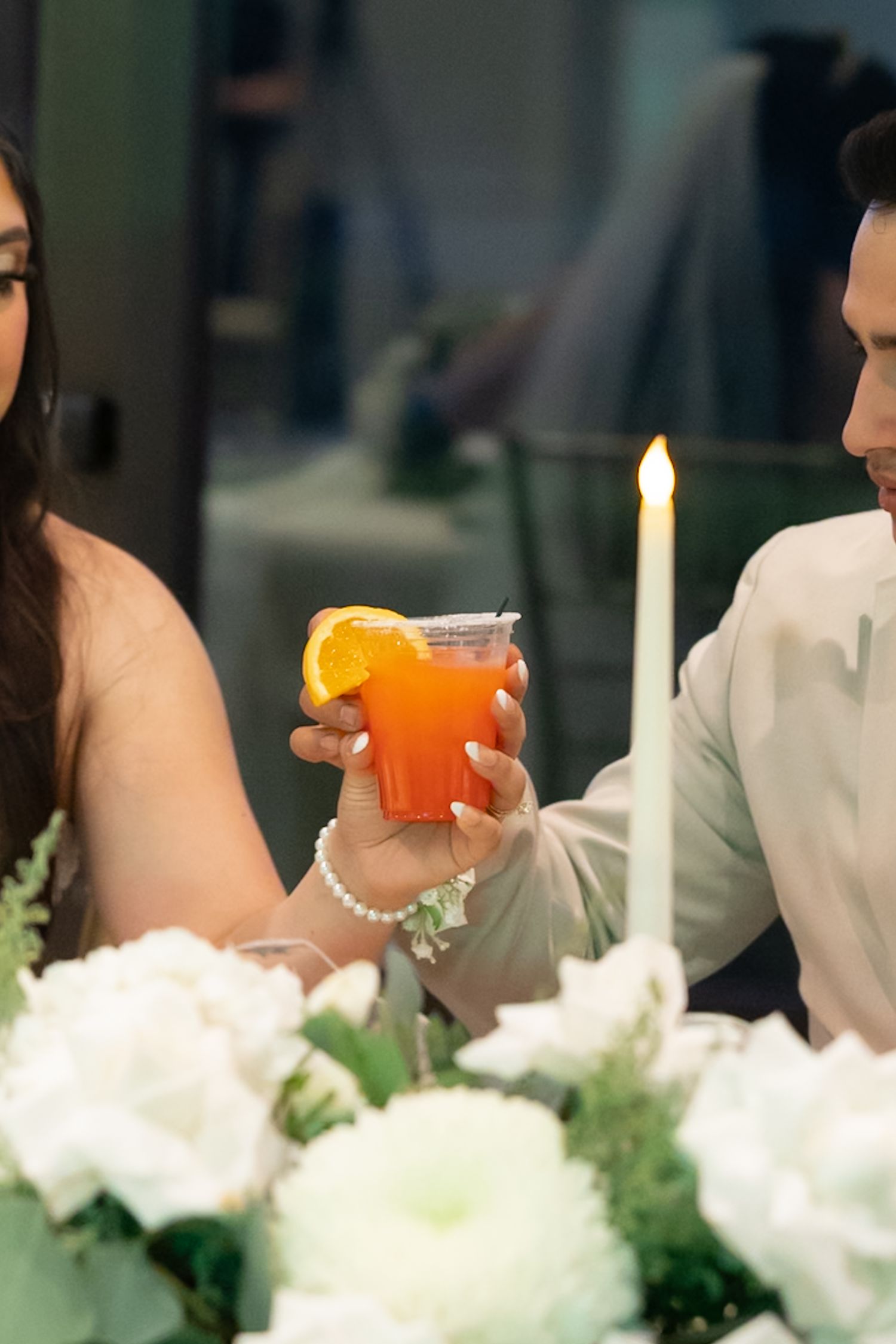 Couple sharing an orange cocktail garnished with a slice of orange at a candlelit table with white flowers.