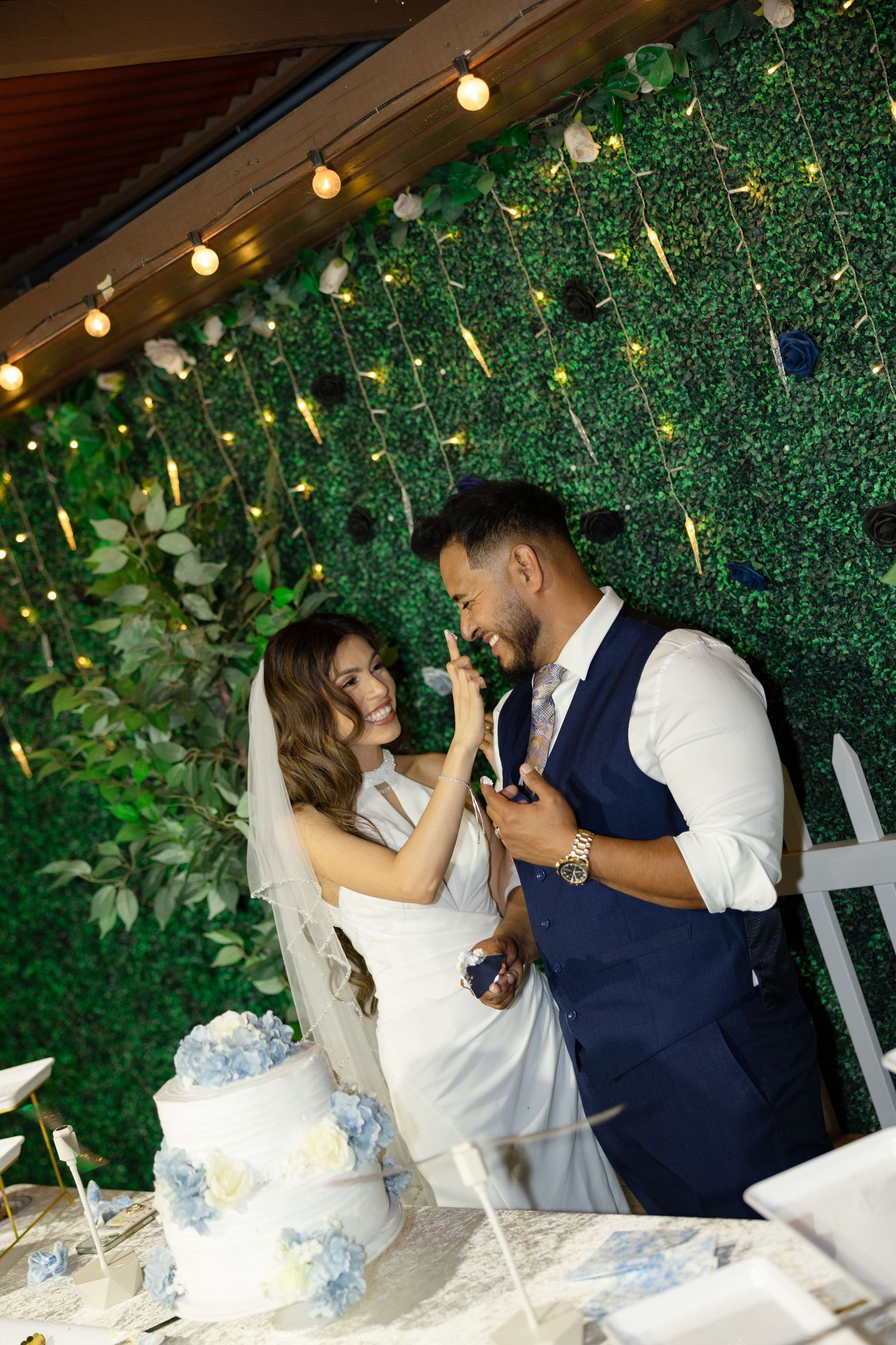 Bride and groom cutting wedding cake at elegant venue with green floral wall and string lights backdrop.