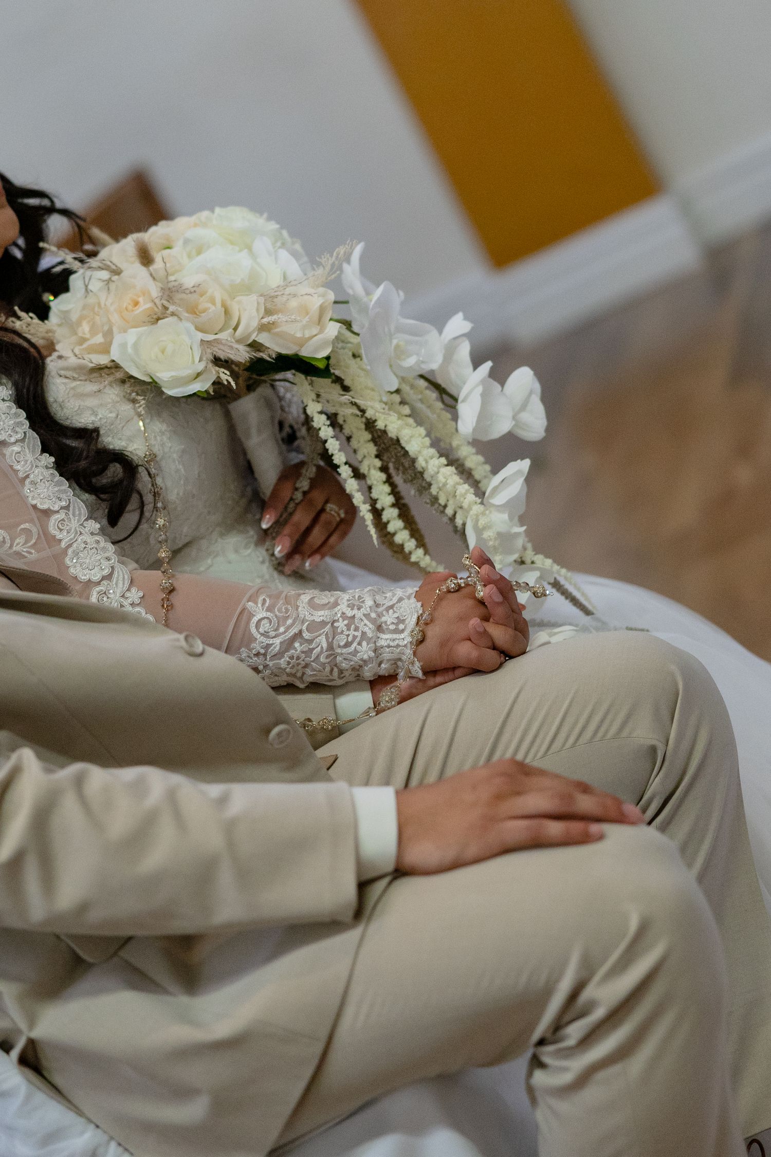 Bride and groom sitting together, bride holding white rose bouquet with pearl accents, groom in beige suit.