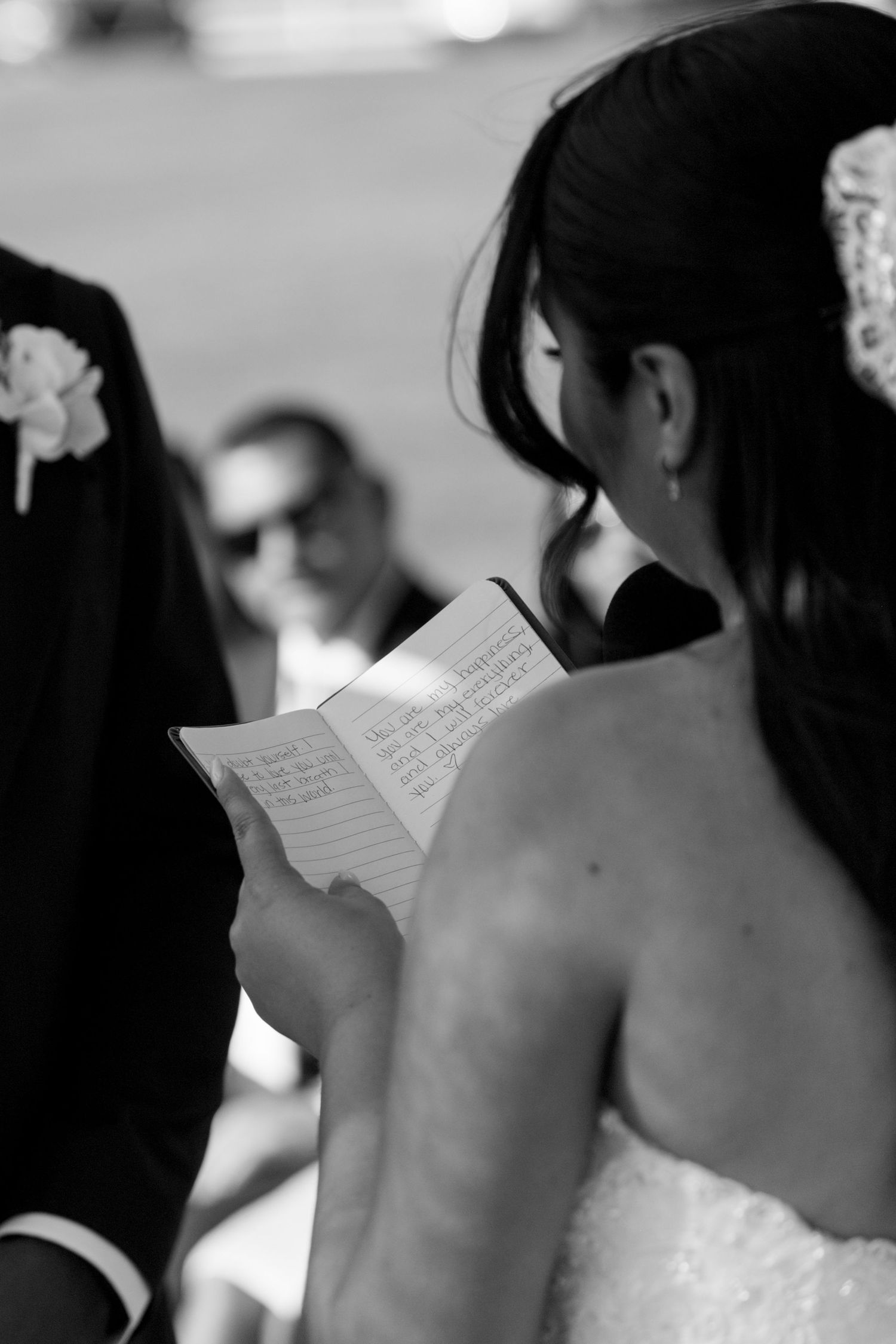 Bride reading handwritten wedding vows from a small notebook during outdoor ceremony in black and white photo.
