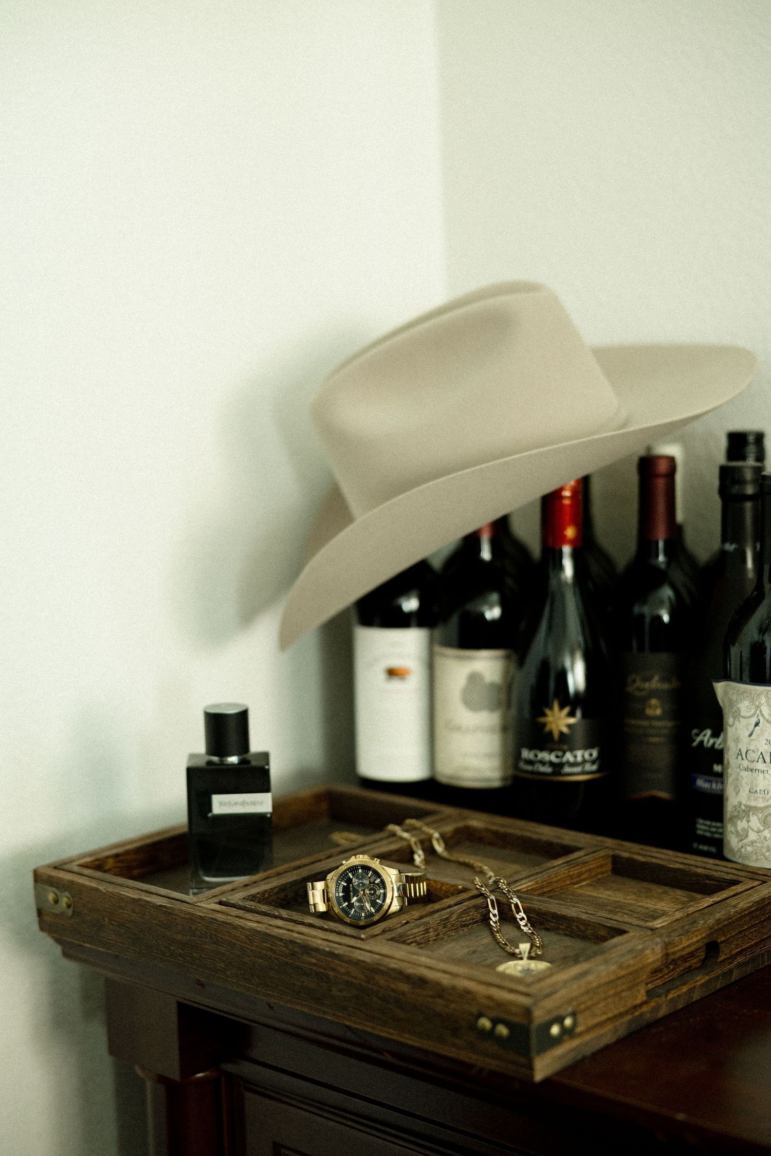 A cowboy hat rests on a wooden tray with a watch and cologne, beside wine bottles on a rustic dresser.