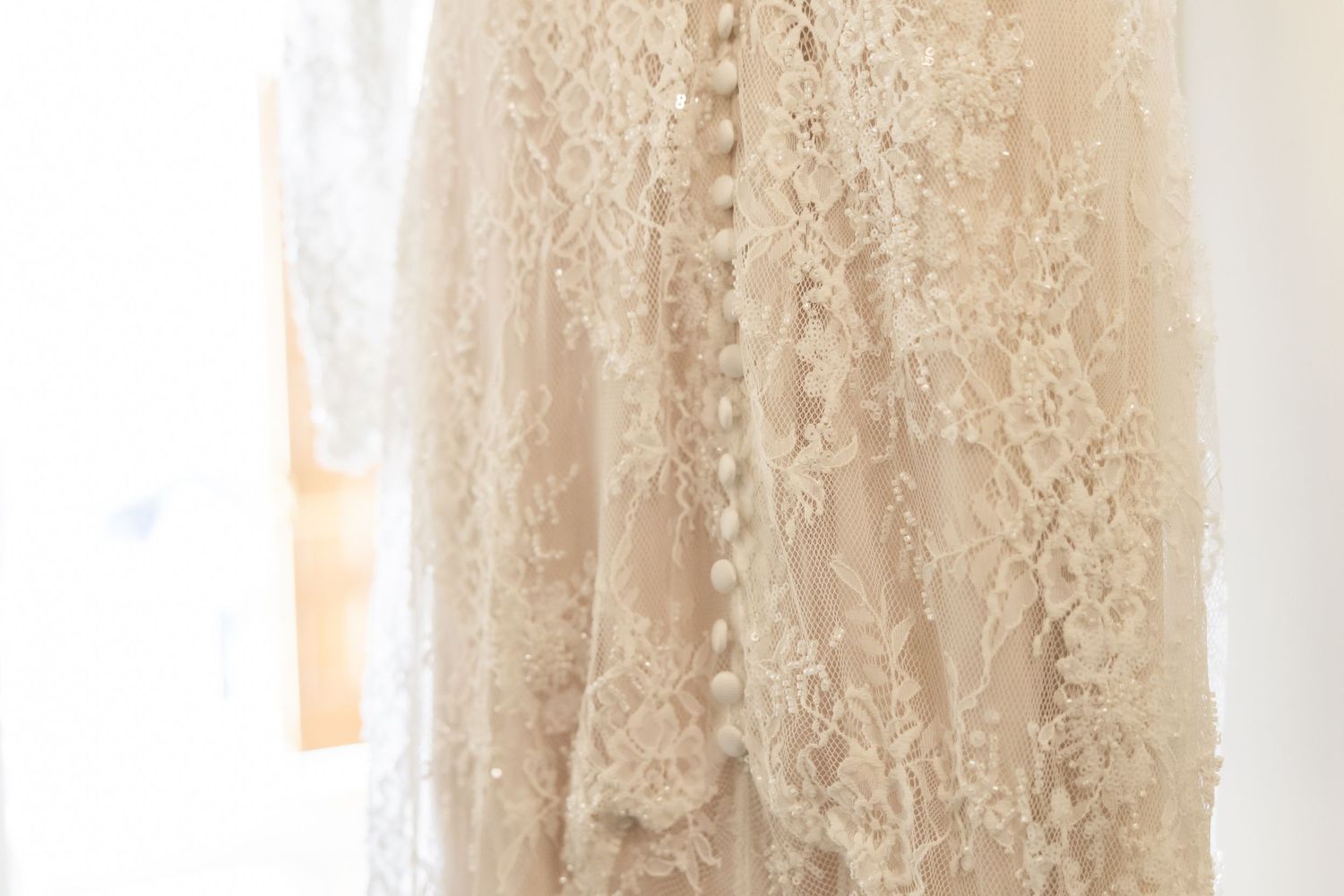 Close-up of an elegant ivory lace wedding dress with delicate floral patterns and button back detail hanging near a window.