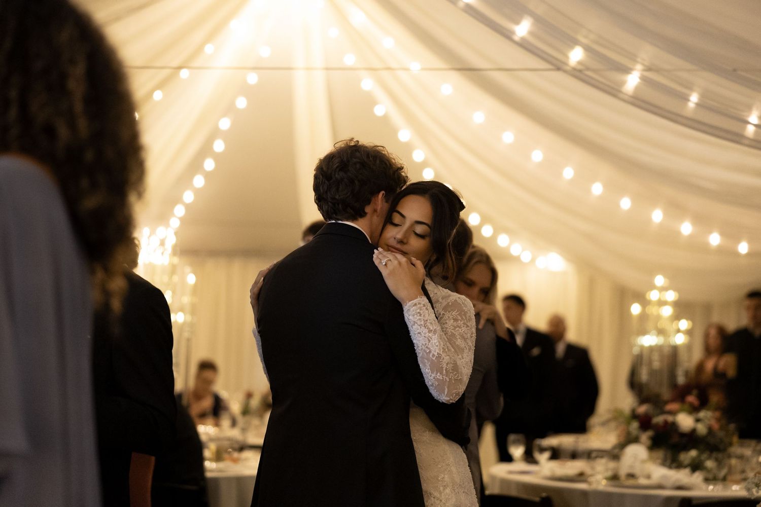 Bride and groom sharing romantic first dance under warm string lights in draped reception tent.