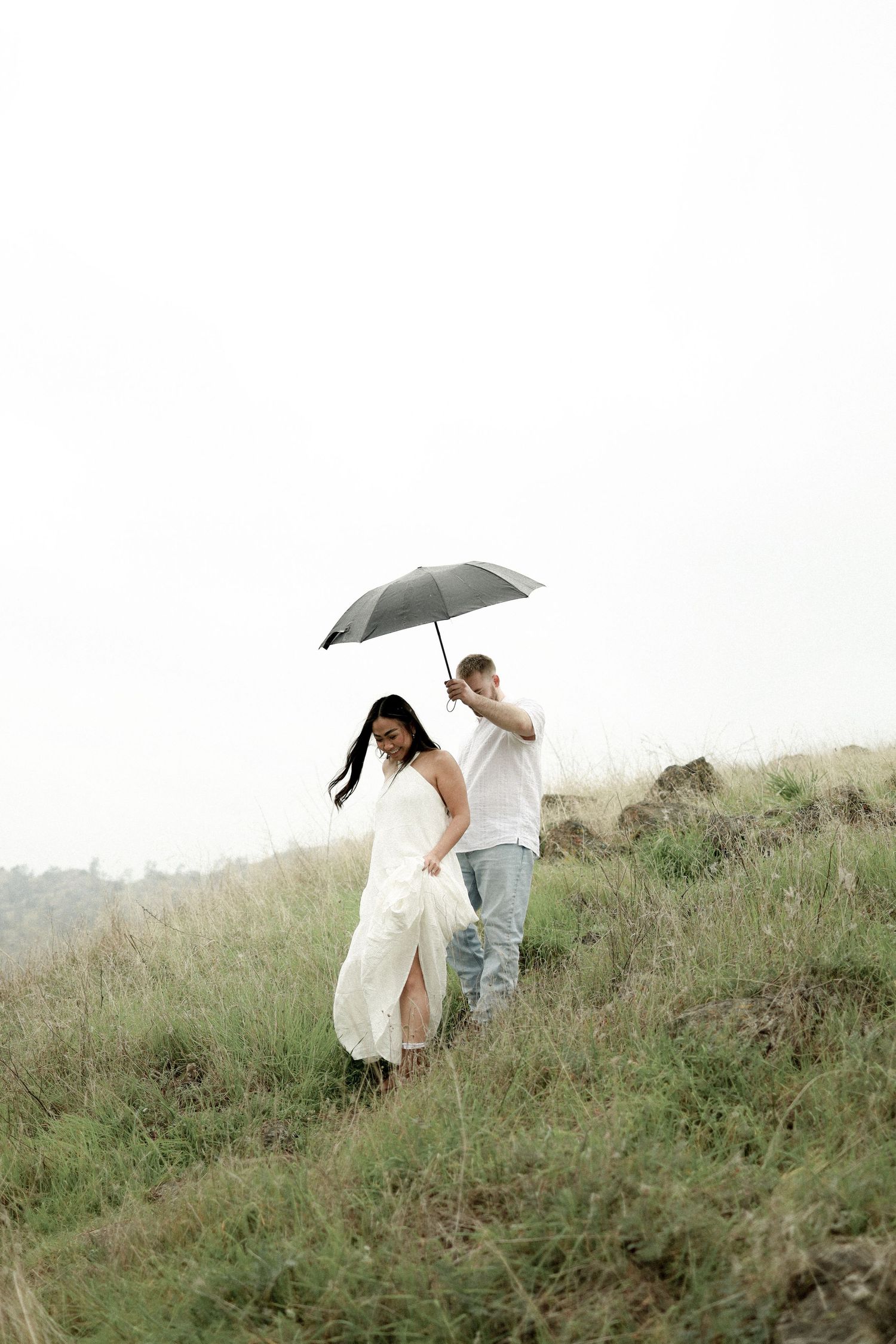 A romantic couple shares a tender moment in a misty field while one holds a white umbrella on an overcast day.
