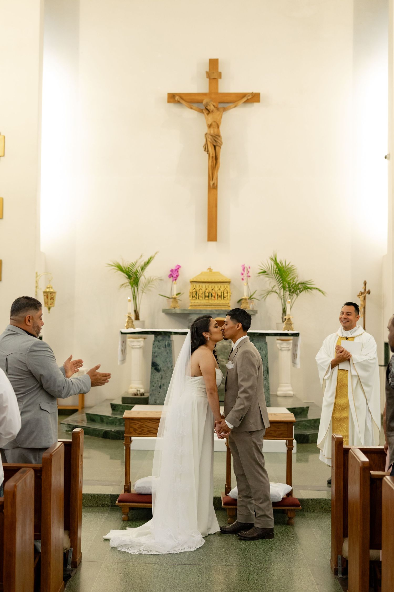 Bride and groom share first kiss at Catholic church altar during wedding ceremony with priest and crucifix in background.