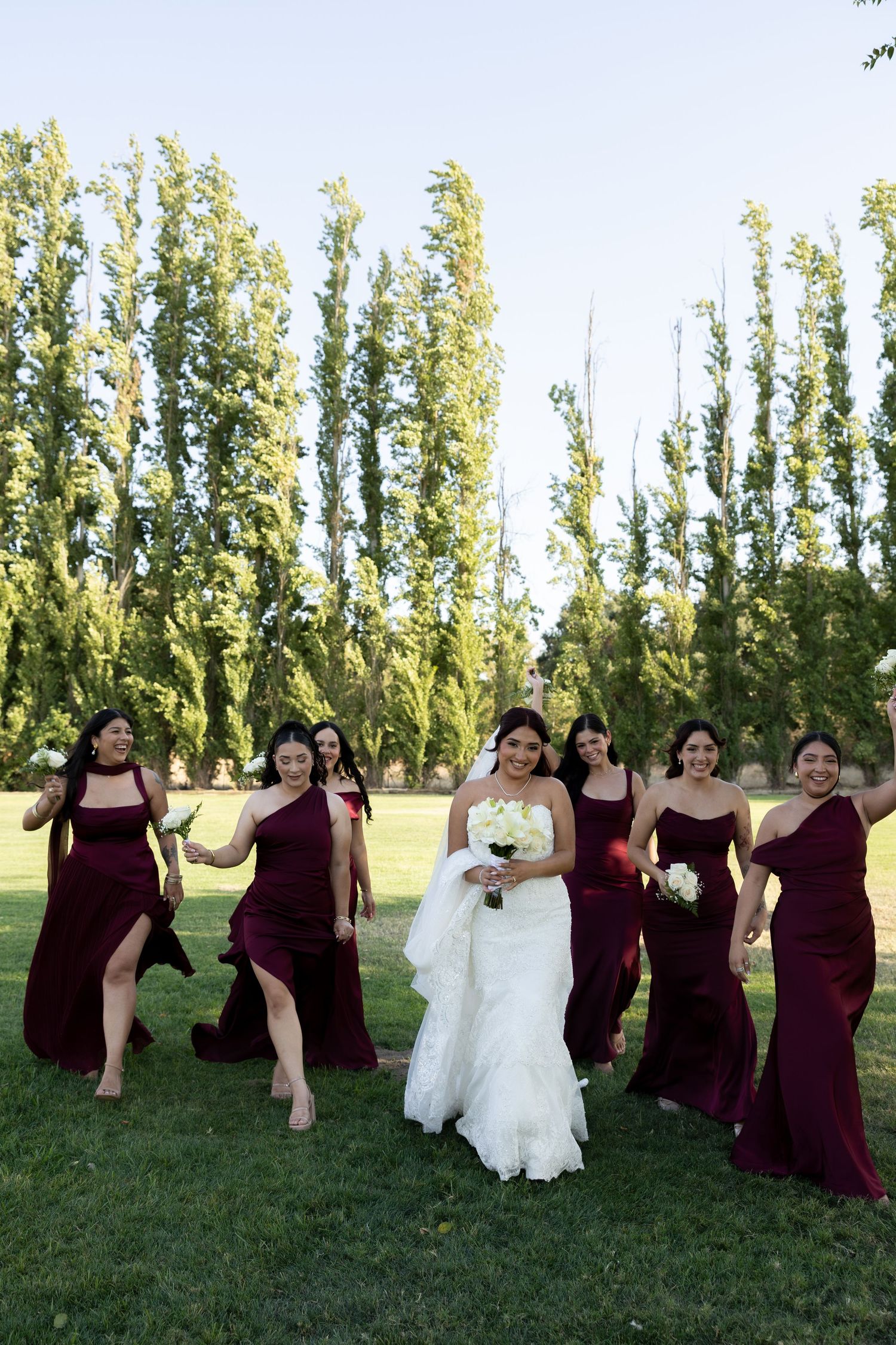 Bride in white lace gown walks with bridesmaids in burgundy dresses carrying bouquets on green lawn.