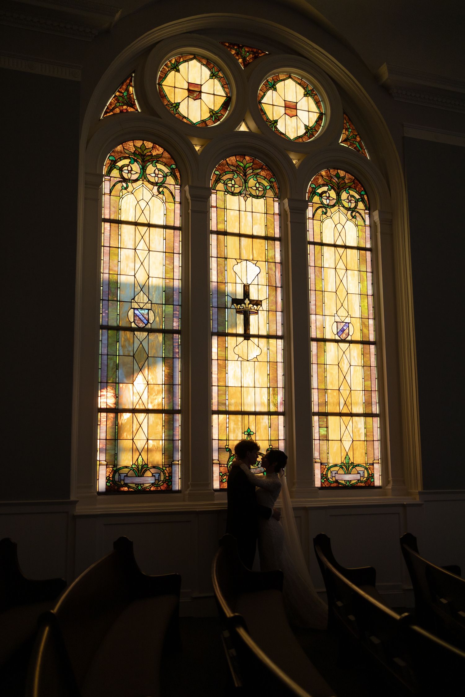 Bride and groom silhouetted before stunning stained glass church windows during wedding.