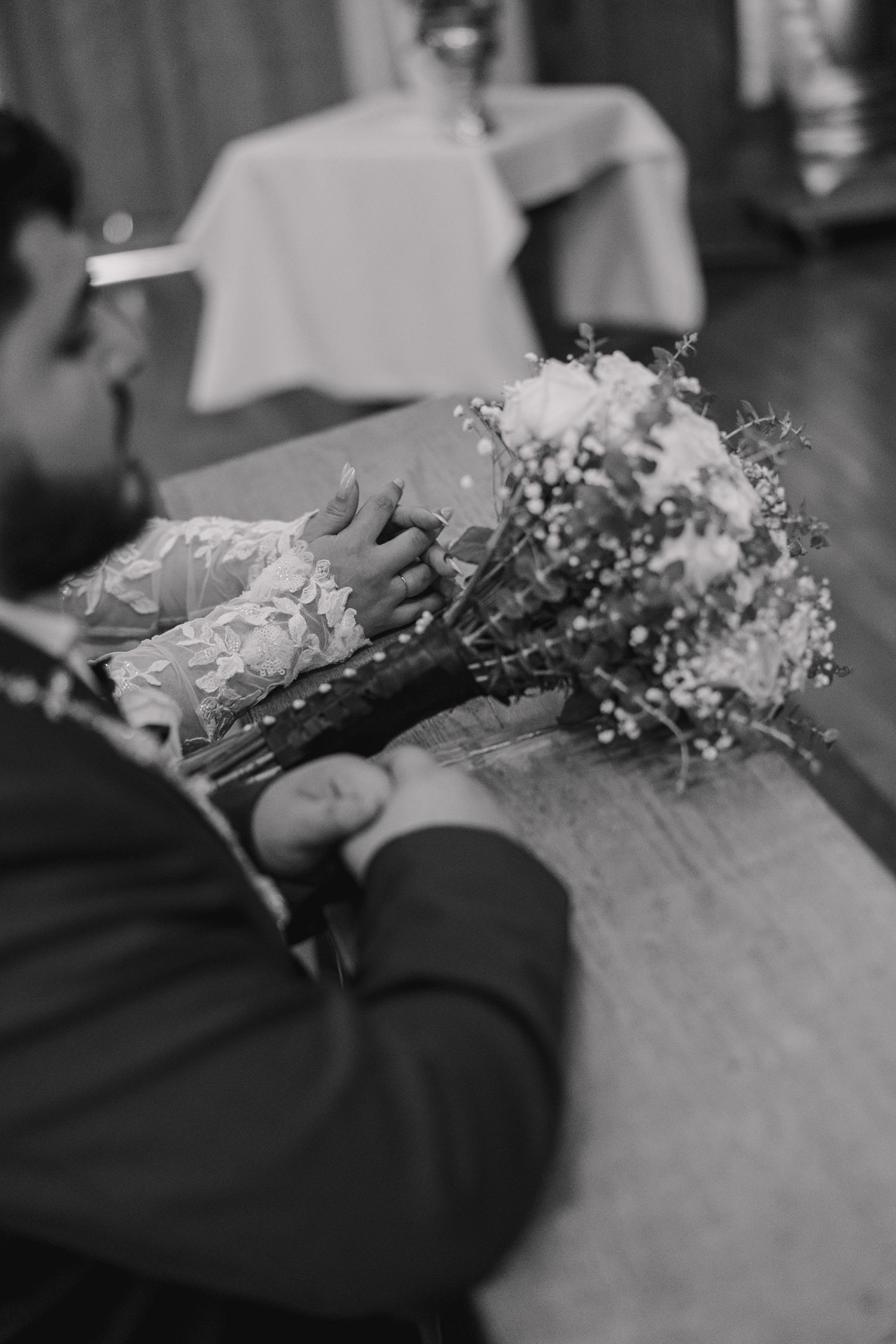 Black and white wedding photo of couple holding hands over a floral bouquet on a wooden table.