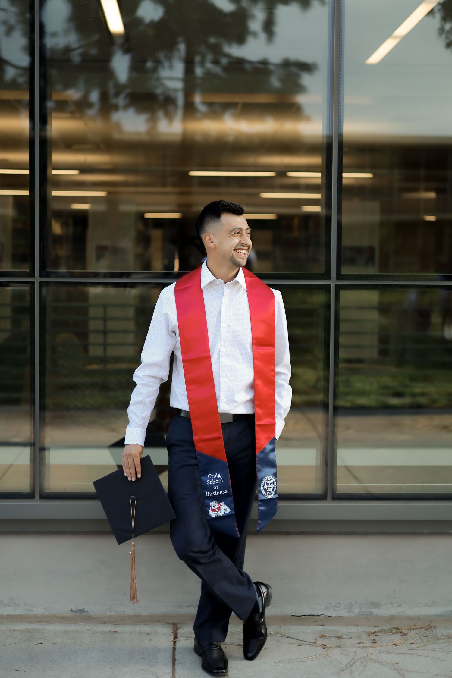 Graduate in white shirt and red stole sits thoughtfully on concrete steps outside modern building.