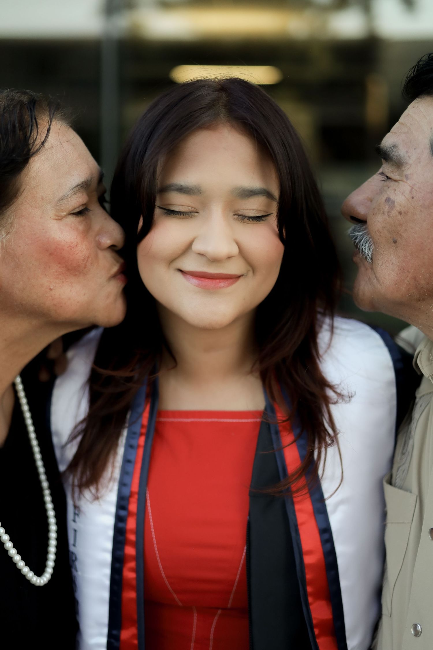 A person in a red top and black vest smiles while others lean in to kiss their cheeks from both sides.