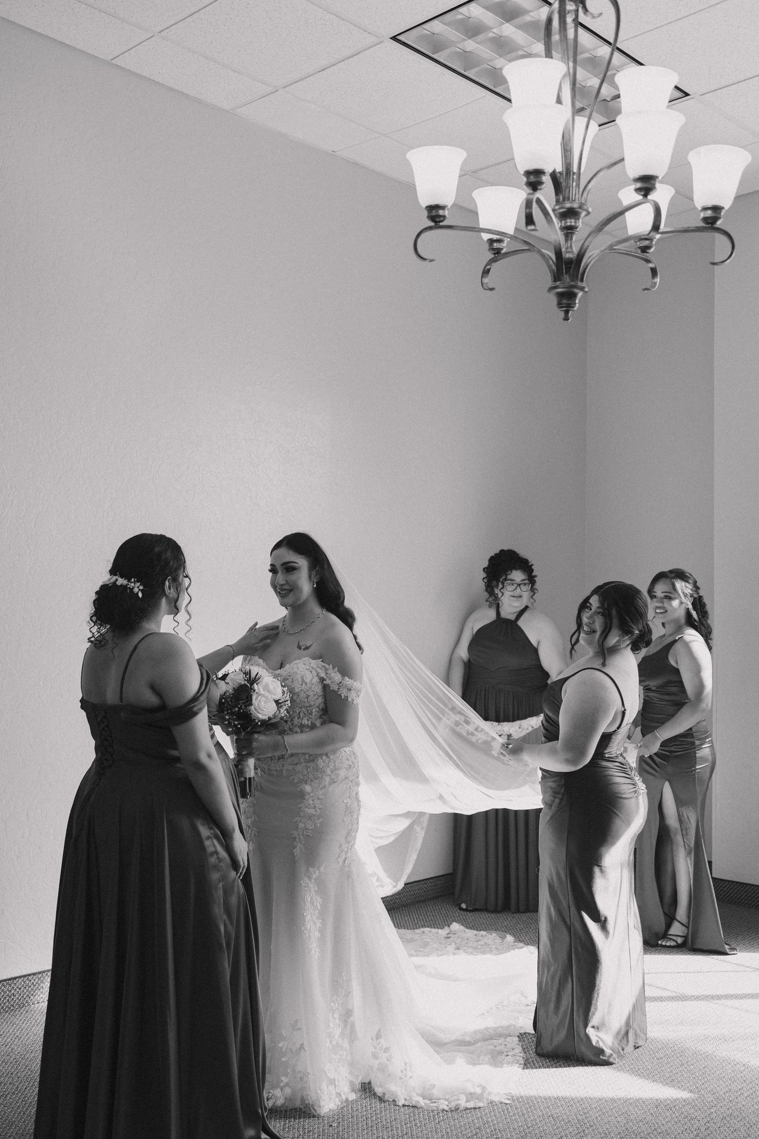 Wedding party helps adjust a flowing white wedding dress in an elegant indoor setting with chandeliers.