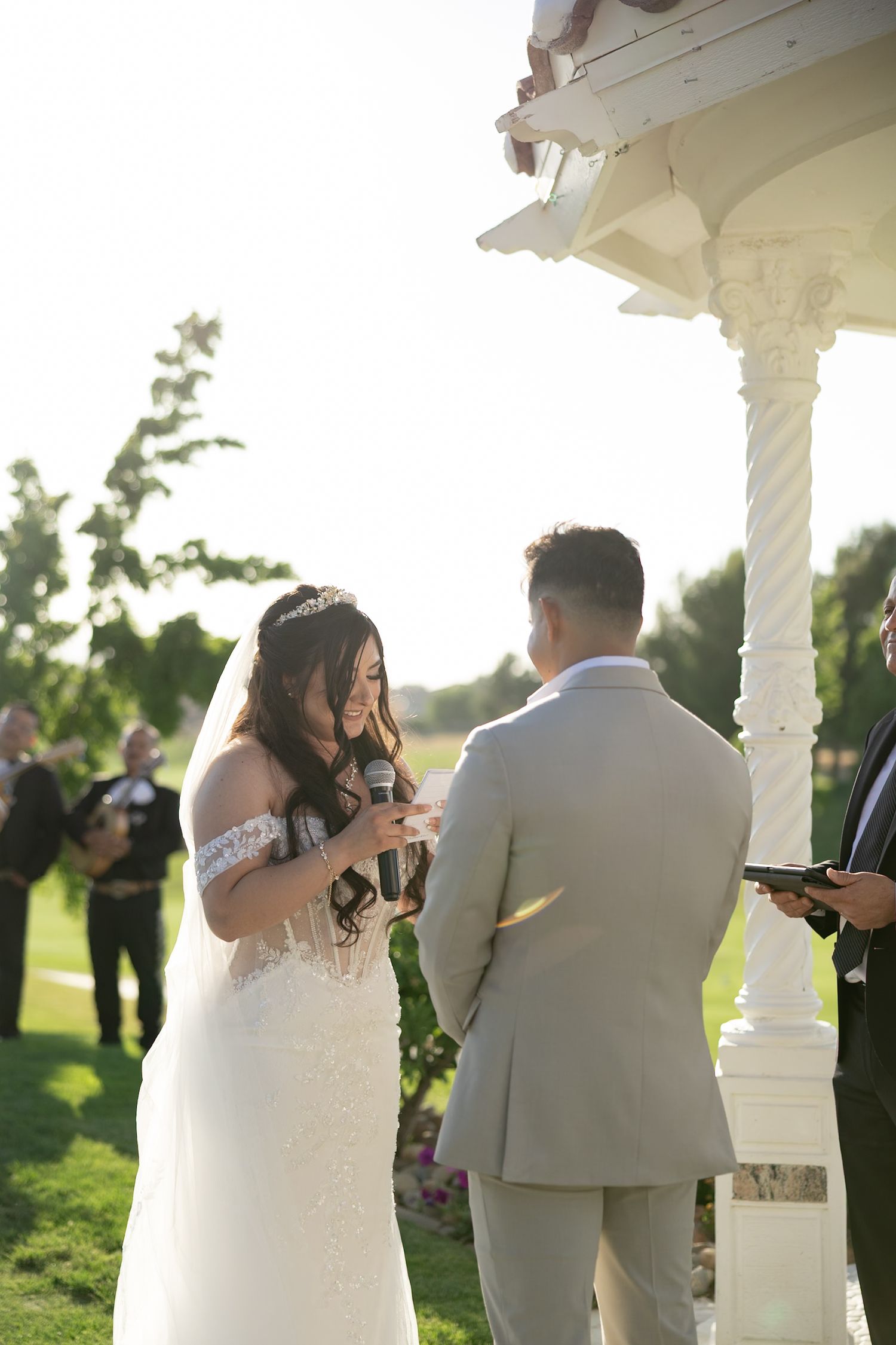 Bride reads heartfelt vows to groom at outdoor gazebo ceremony surrounded by lush greenery in golden sunlight.