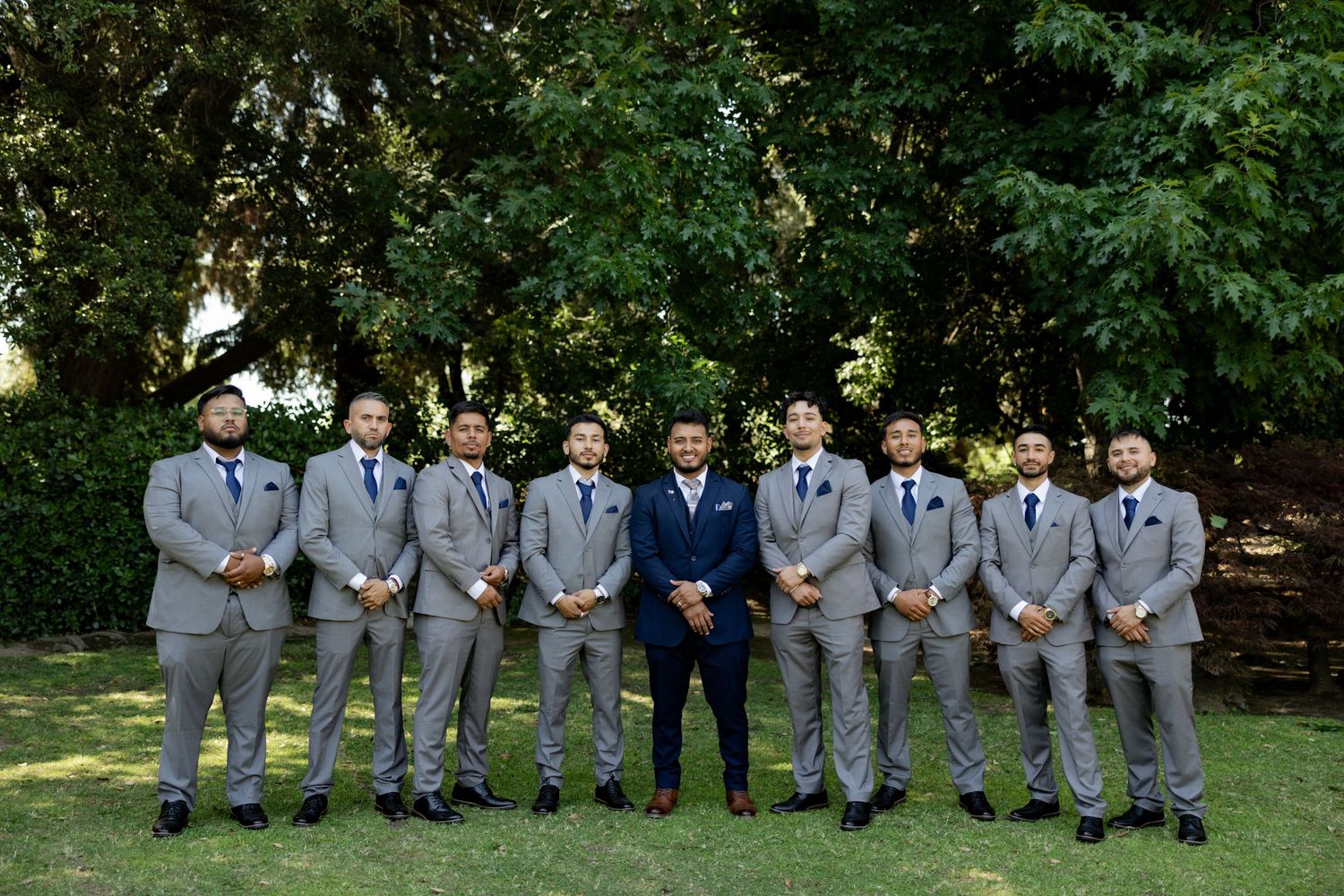 Groom in navy suit stands with nine groomsmen in grey suits and blue ties outdoors on green lawn with trees.