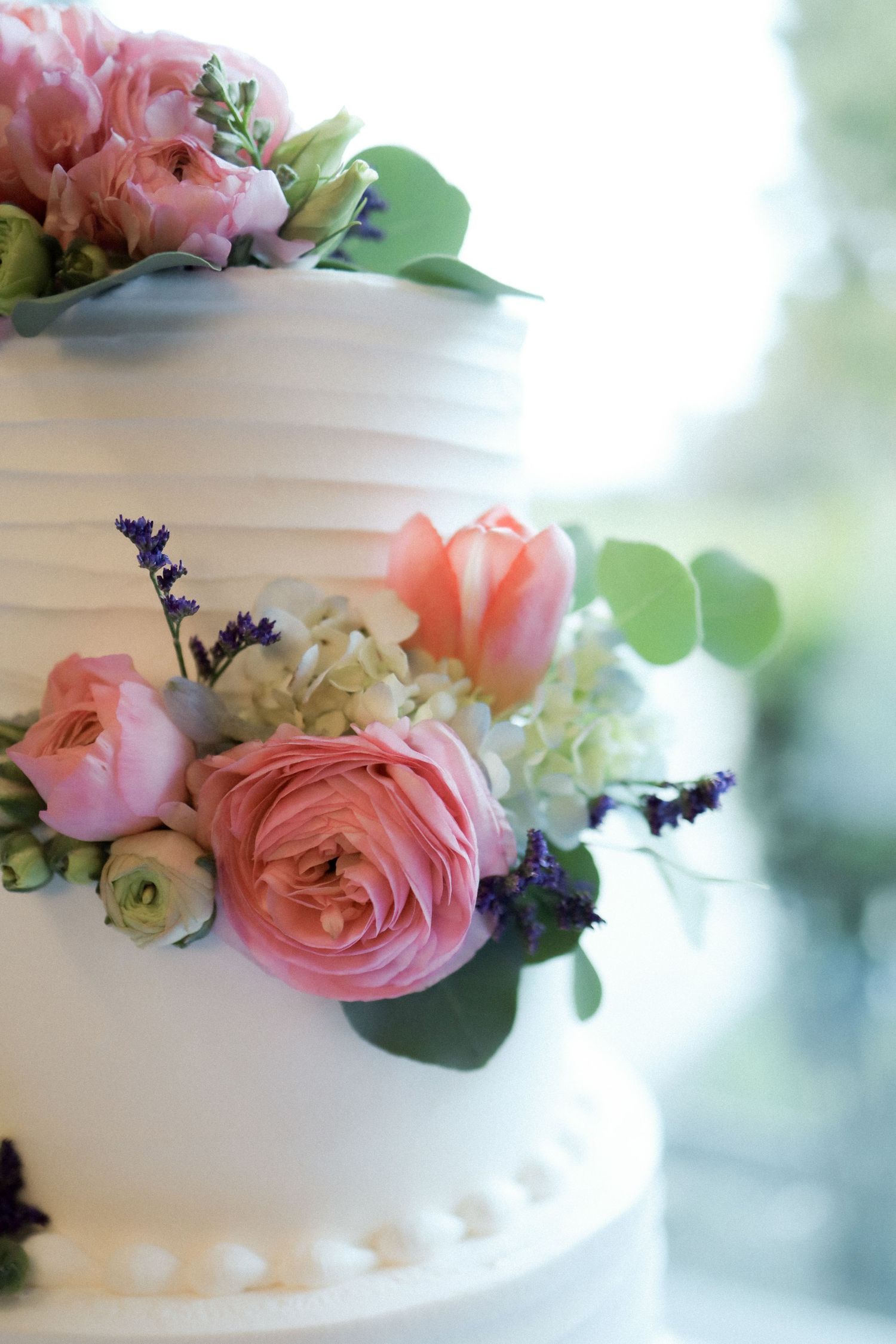 White wedding cake decorated with delicate pink roses and lavender flowers against a soft blurred background.