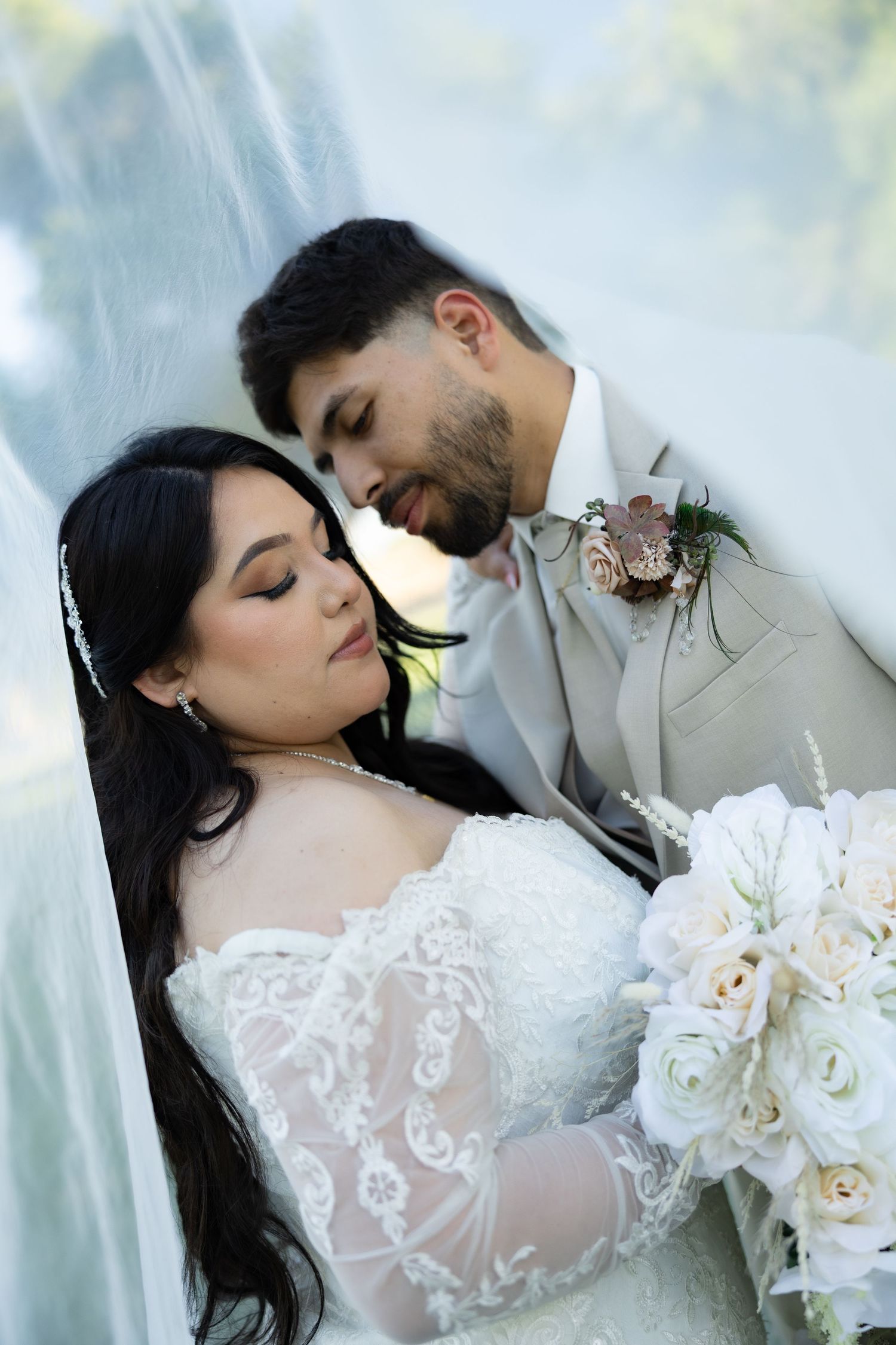 A bride and groom in elegant wedding attire share a tender moment, she holds white roses bouquet, he wears a gray suit with boutonniere.