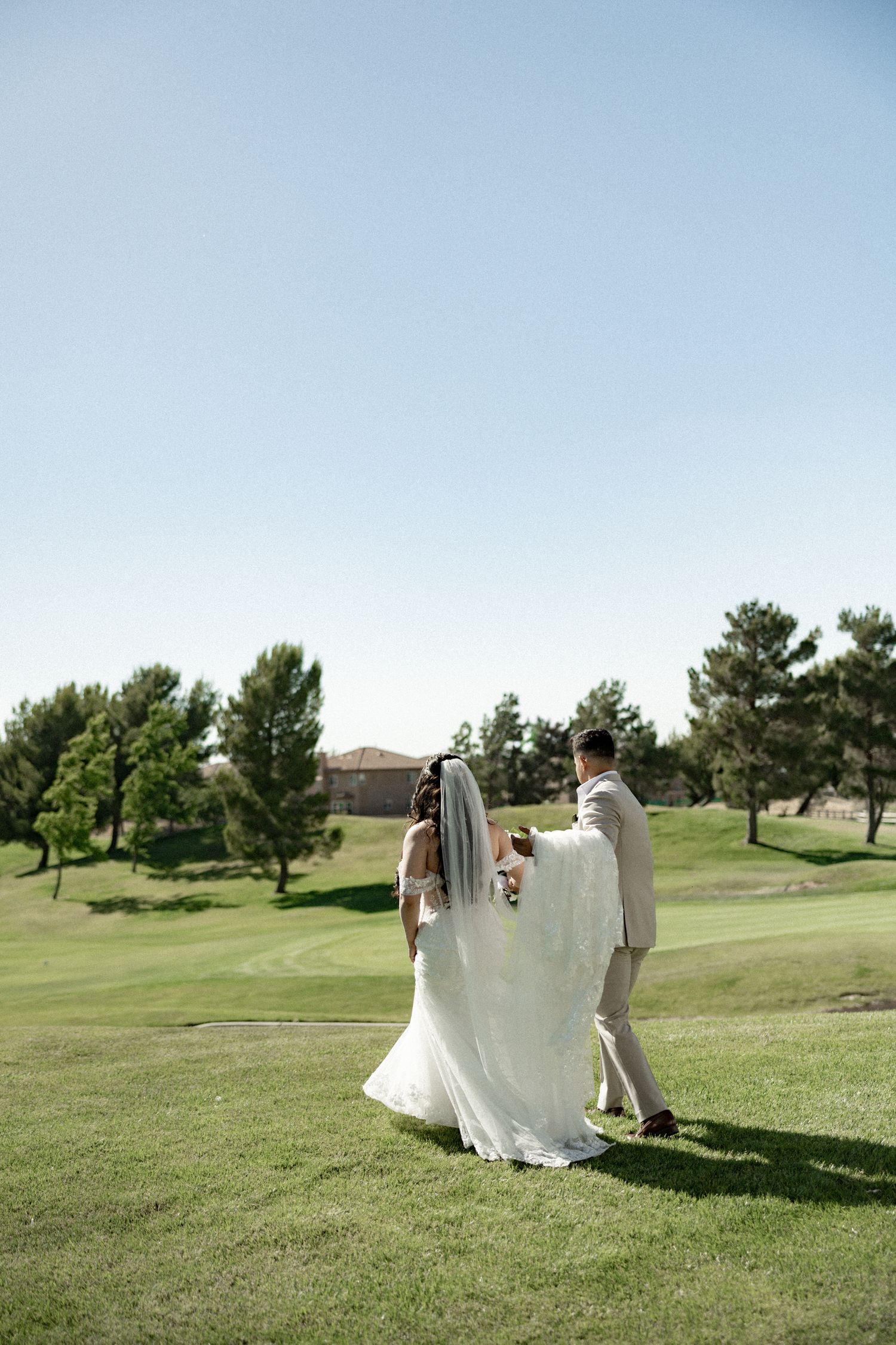 Bride in flowing white gown and veil walks with groom across green golf course under clear sky.